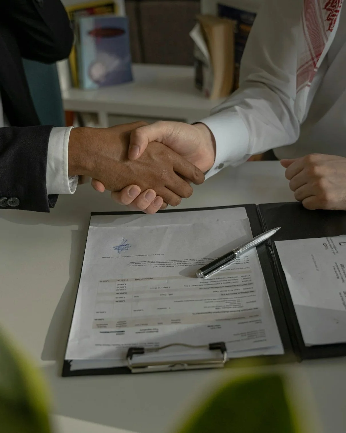 Two individuals shake hands over a desk with documents and a pen, indicating a formal agreement or business meeting.