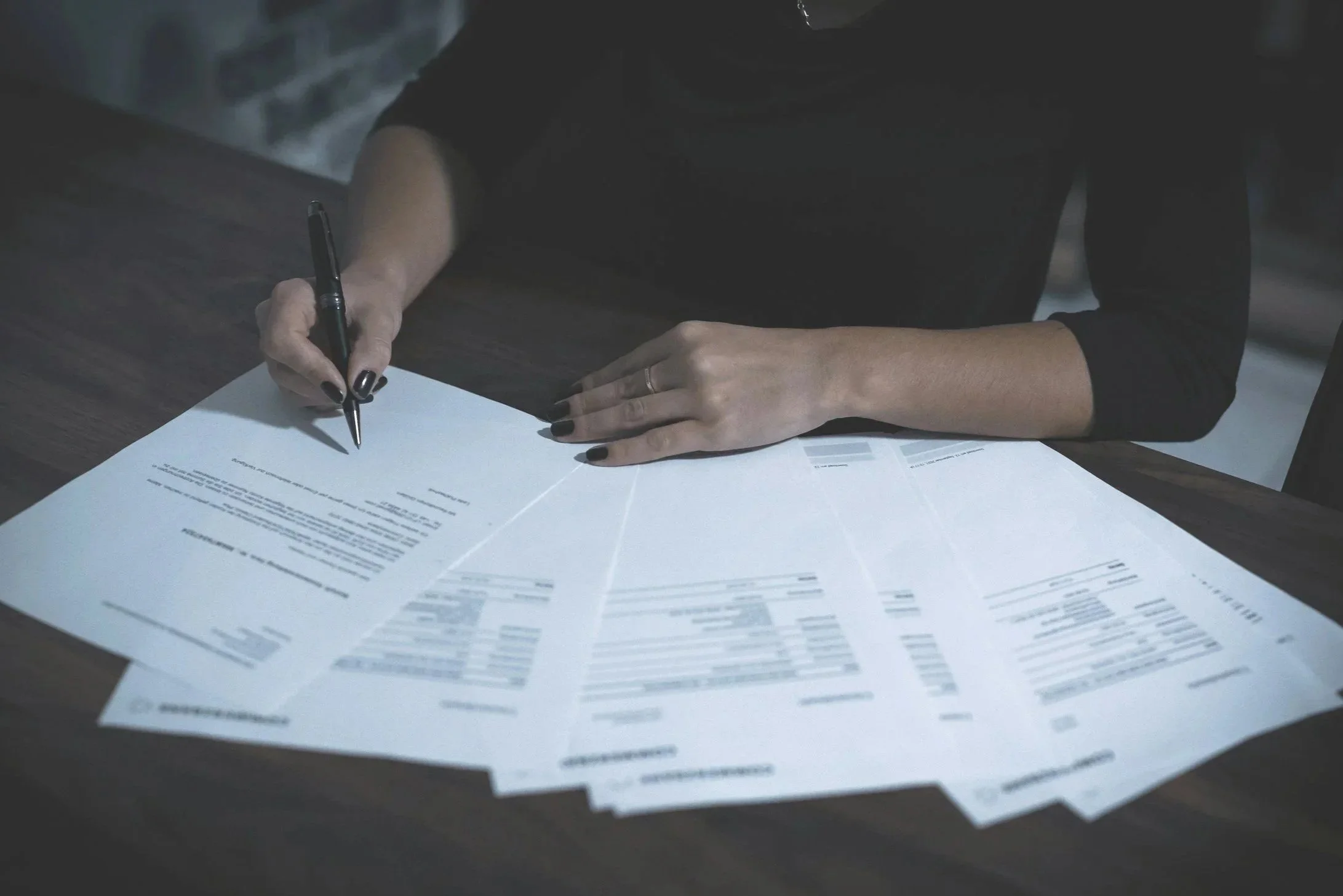 Person in black clothing writing or signing documents on a dark wooden table with scattered papers.