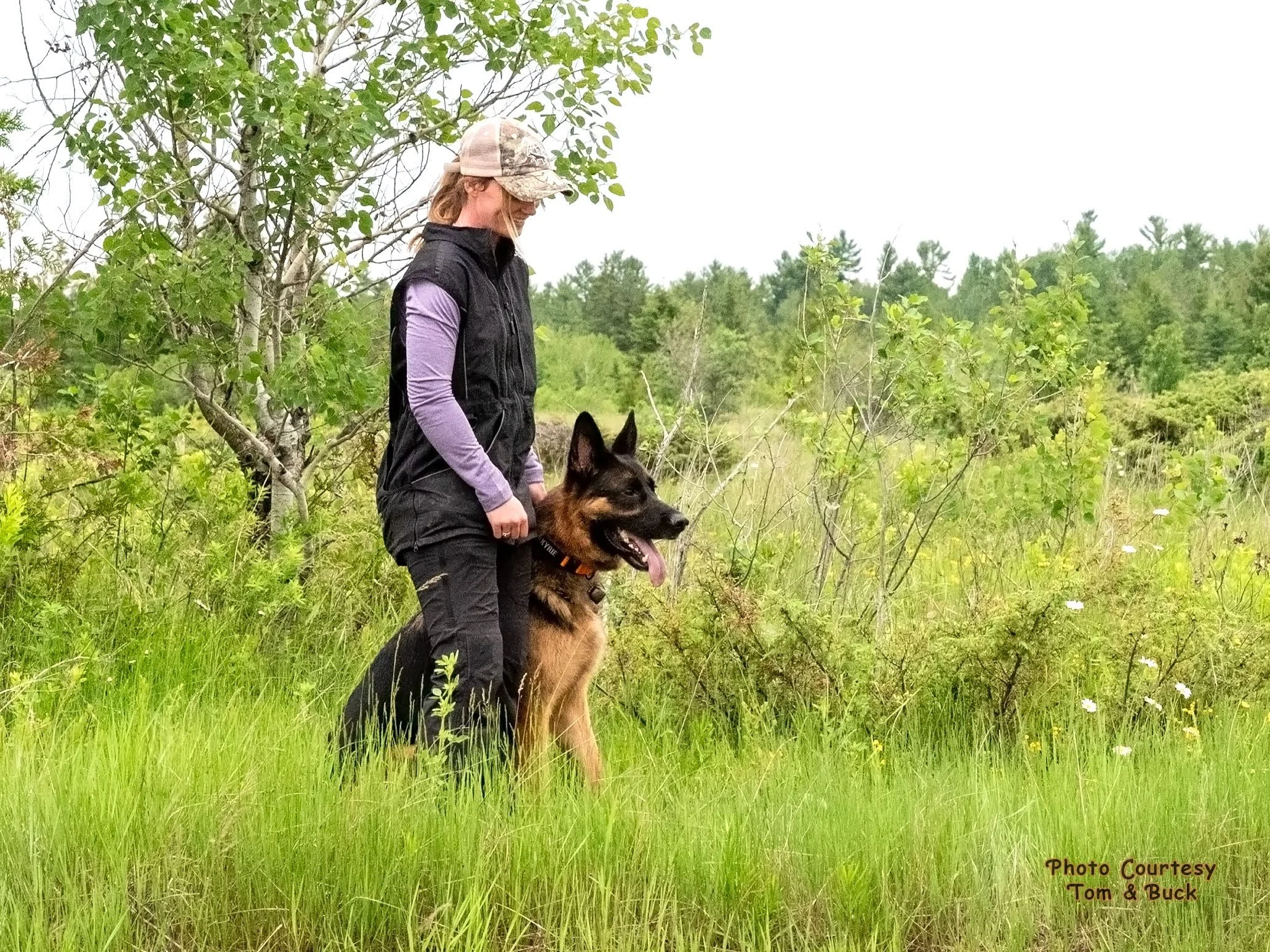 A woman in outdoor clothing and a camouflage baseball cap stands with a German Shepherd dog in a green, grassy, wooded area during daytime.