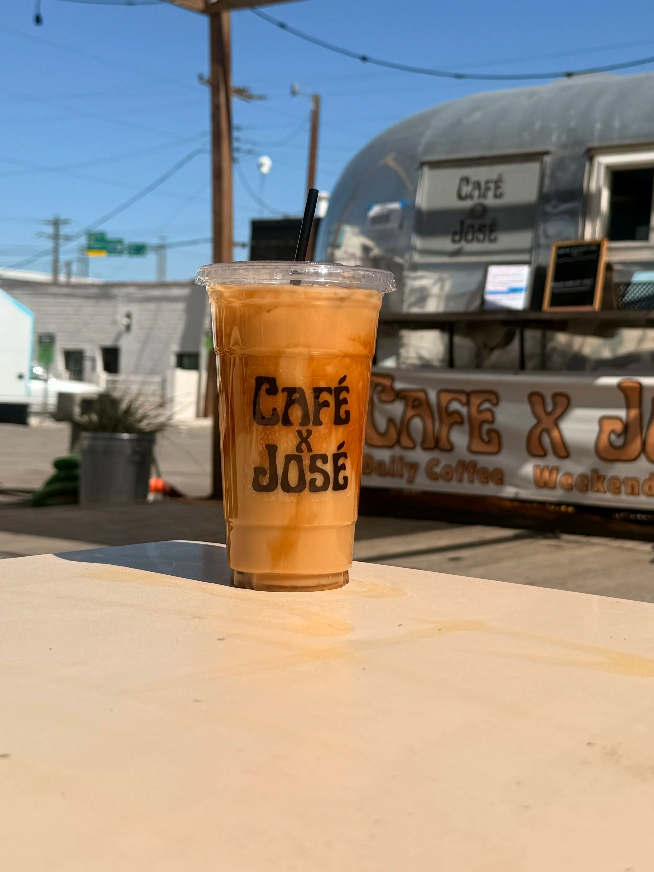 A plastic cup filled with iced coffee or tea on a beige table outdoors, with a food truck or mobile cafe in the background. The cup has a black straw and the words 'Café José' printed on it.