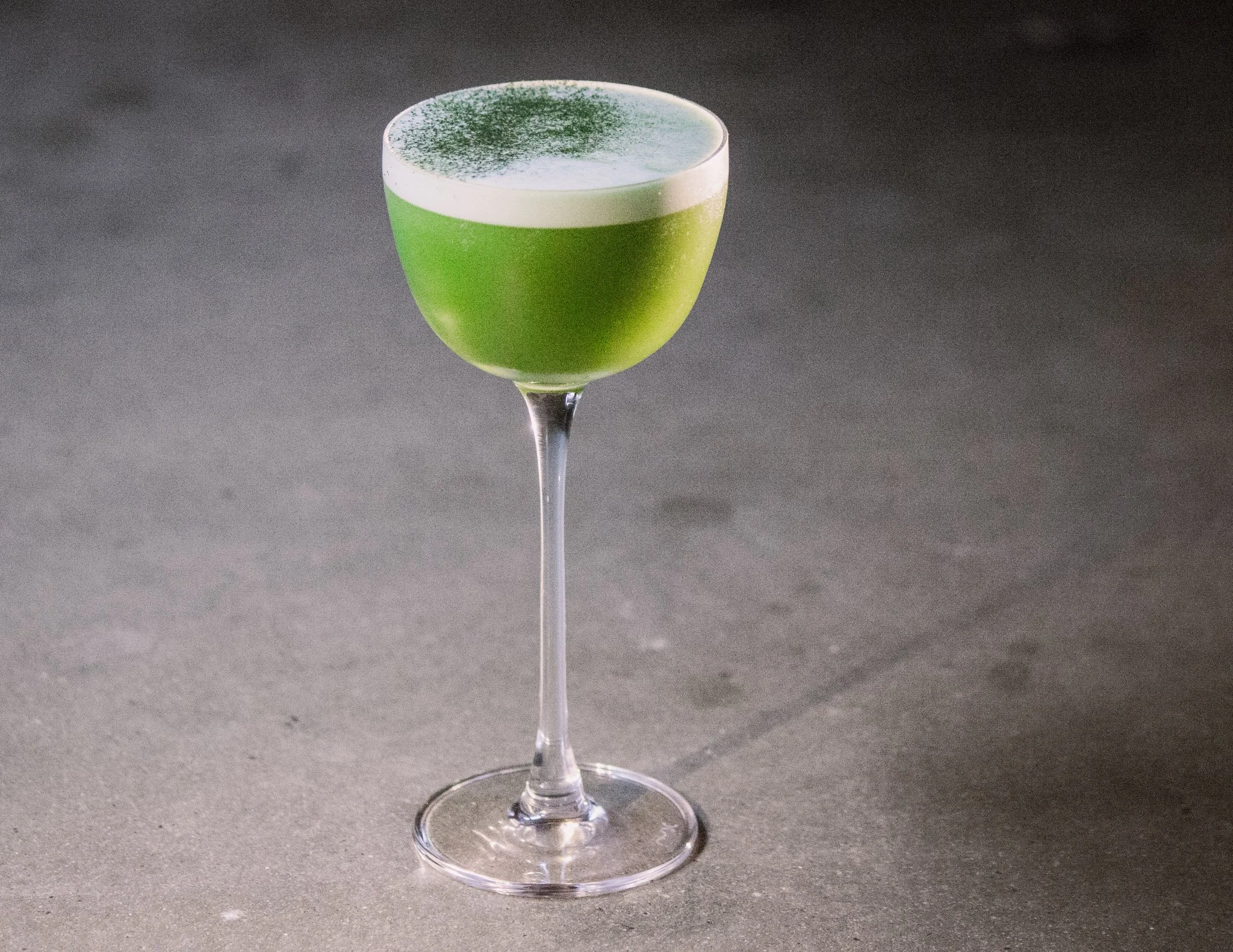 A green cocktail in a coupe glass with a white foam top sprinkled with green powder, placed on a dark surface.