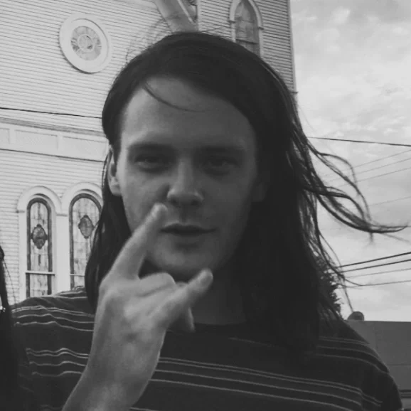 A young man with long, dark hair making a peace sign near his face. He is standing outdoors in front of a building with arched windows and some power lines visible in the background, in black and white.
