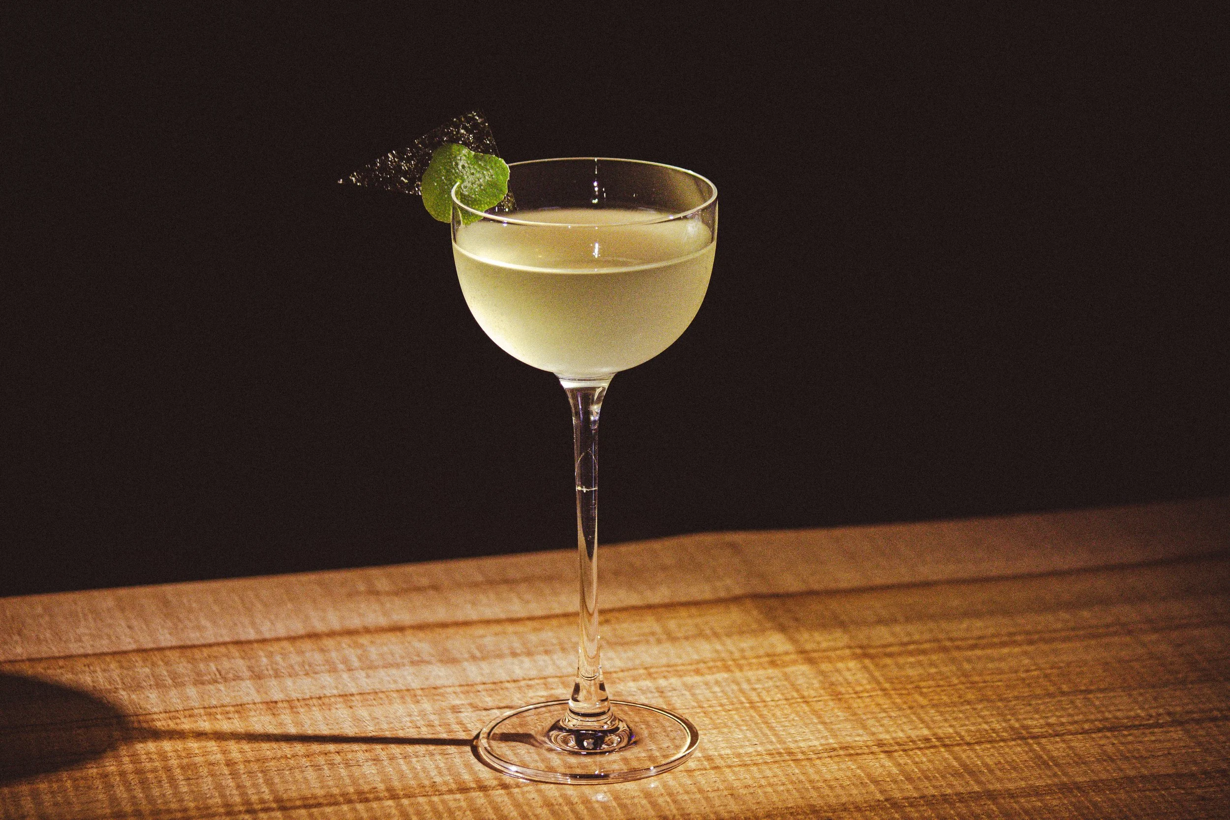 A cocktail glass filled with a pale yellowish cocktail, garnished with a lime wedge, on a wooden surface against a dark background.