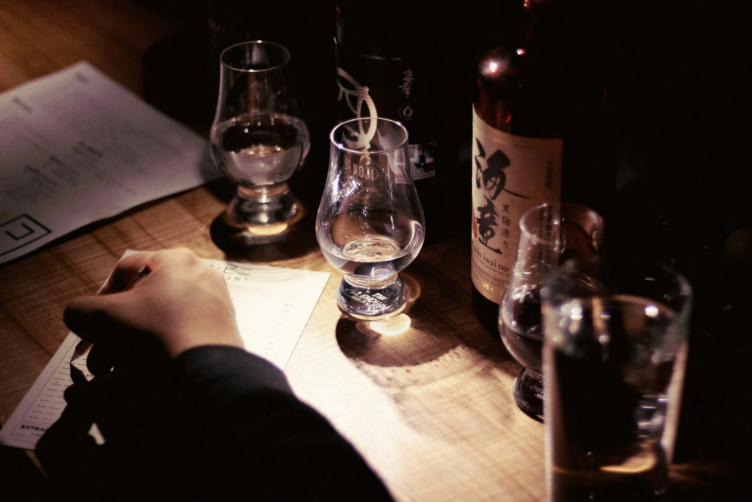 A person sitting at a wooden table with various glasses of water and alcohol bottles, a menu, and a notepad visible in a dimly lit setting.