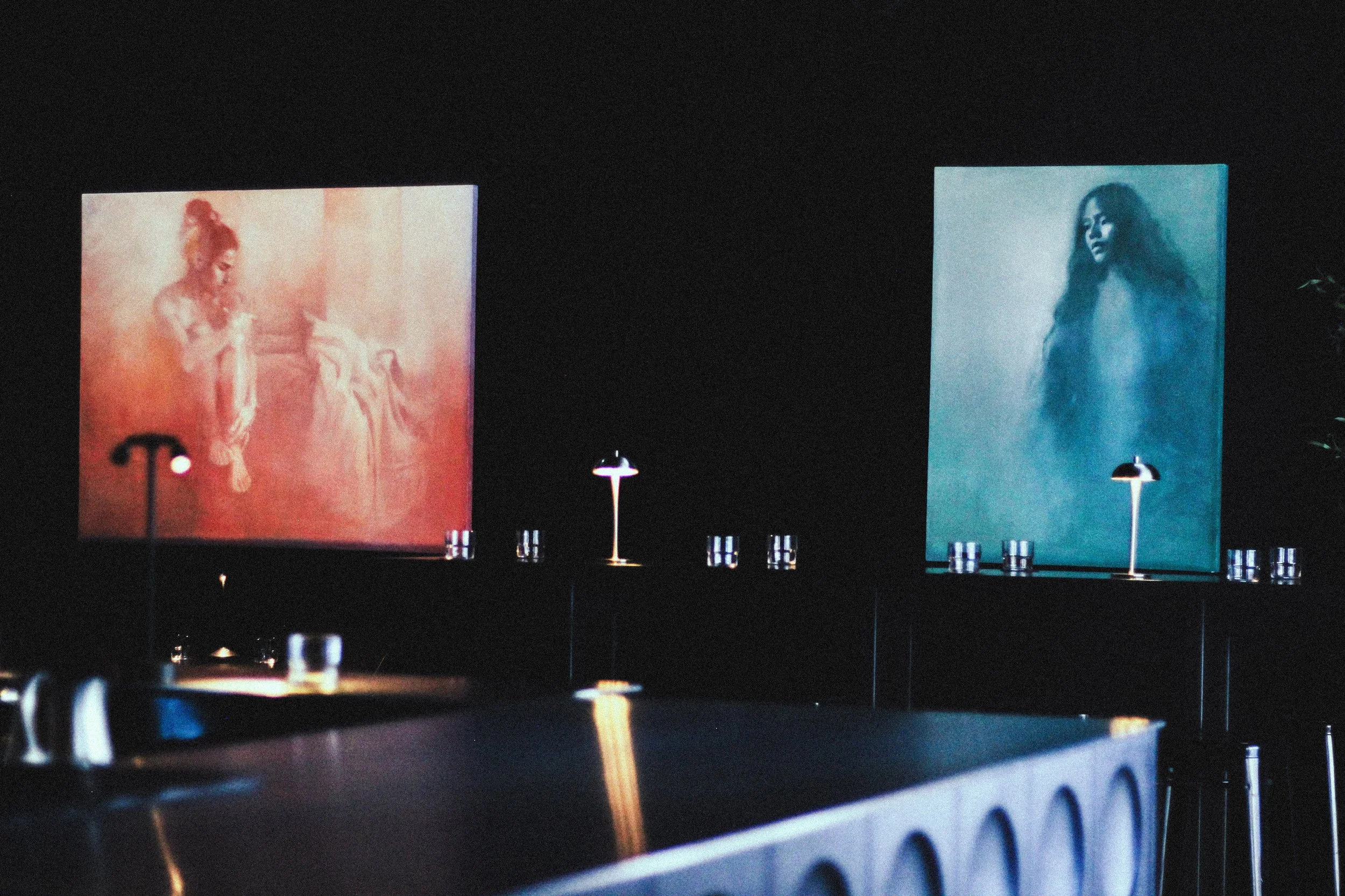 A dark room with two illuminated large portraits on standing boards; one portrait of a young woman with her hair in a bun and the other of a woman with long flowing hair.