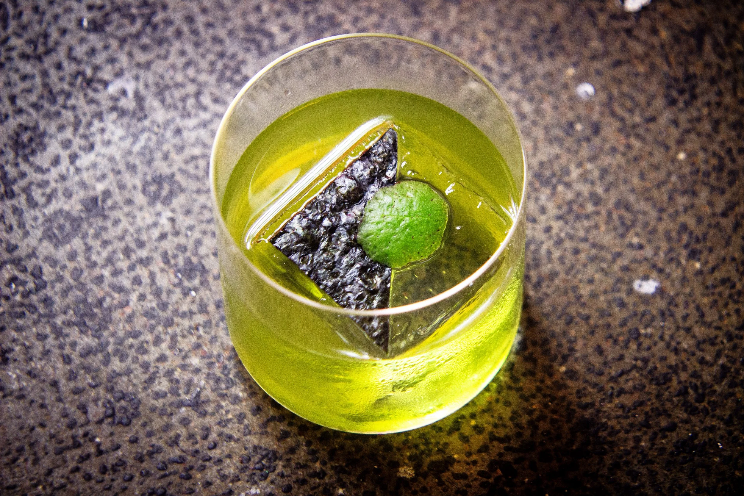 A glass filled with a green drink, ice, a lime wedge, and a piece of seaweed, on a dark speckled countertop.