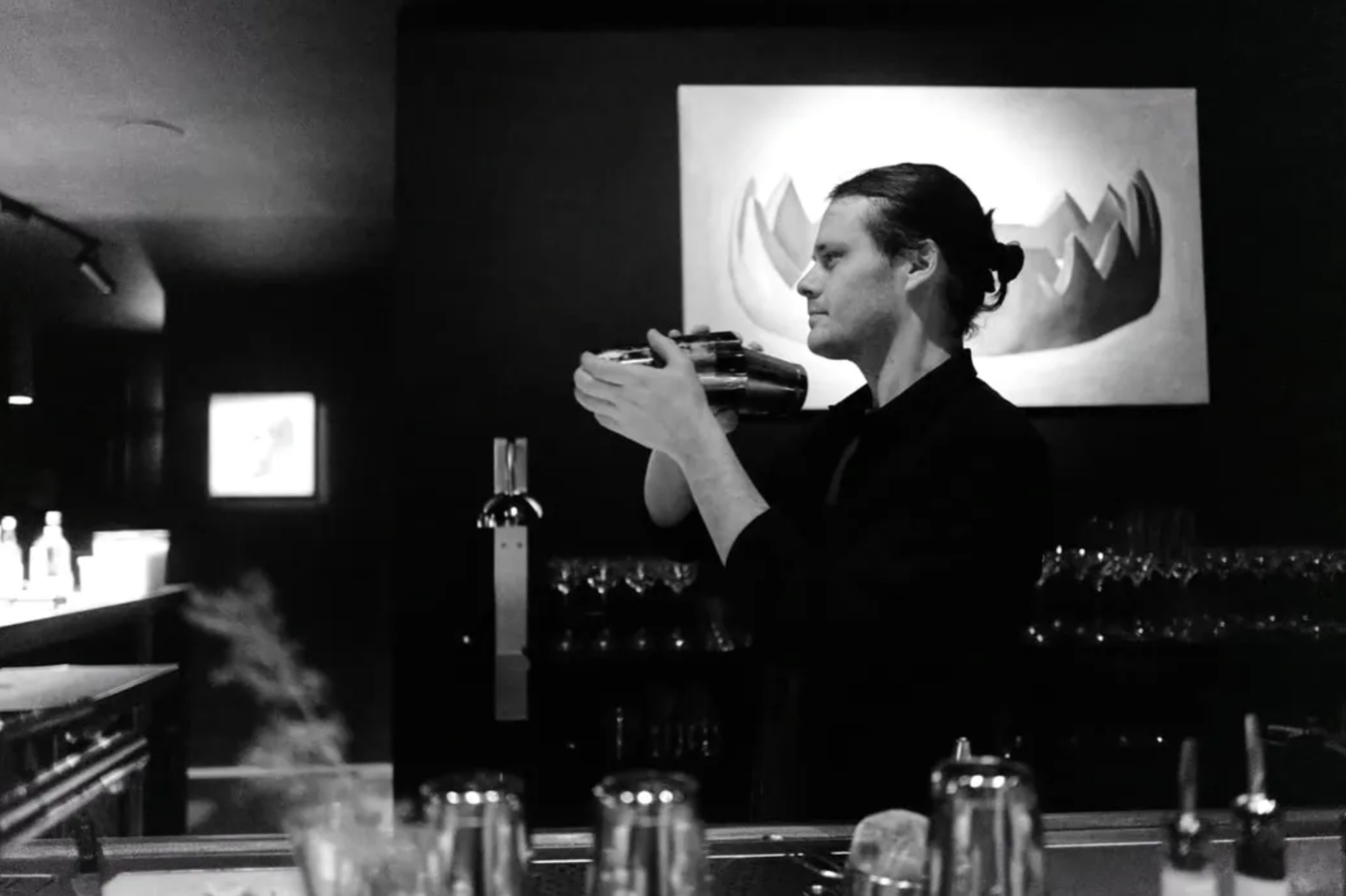 A man with dark hair in a bun, wearing a dark shirt, holds a cocktail glass with an upside-down bottle in a bar or club setting. There is a large framed artwork behind him, lit with bright light, and blurred glasses and bar bottles in the foreground.