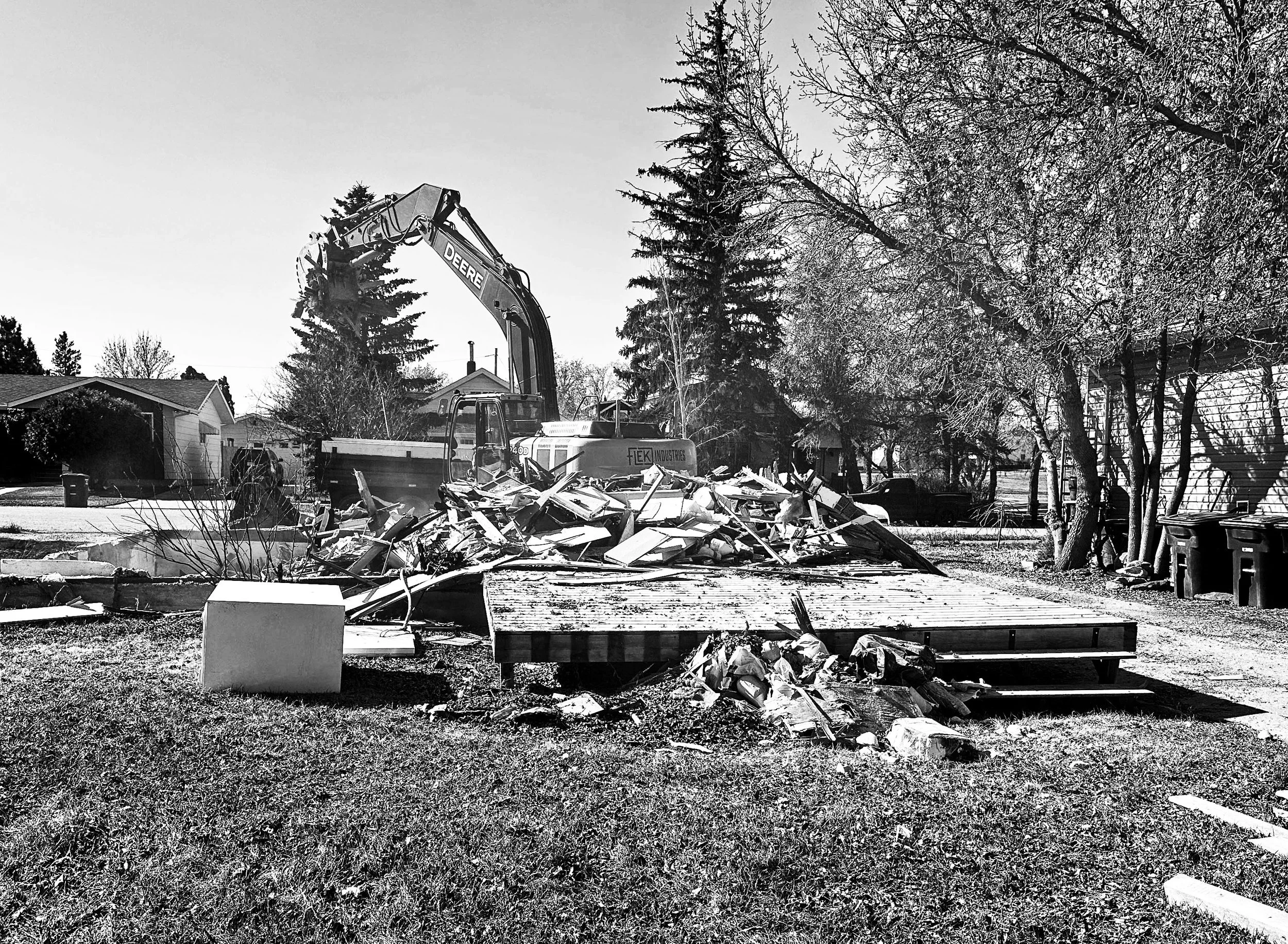 A construction demolition site in a neighborhood with a large excavator and piles of debris, surrounded by leafless trees and houses.