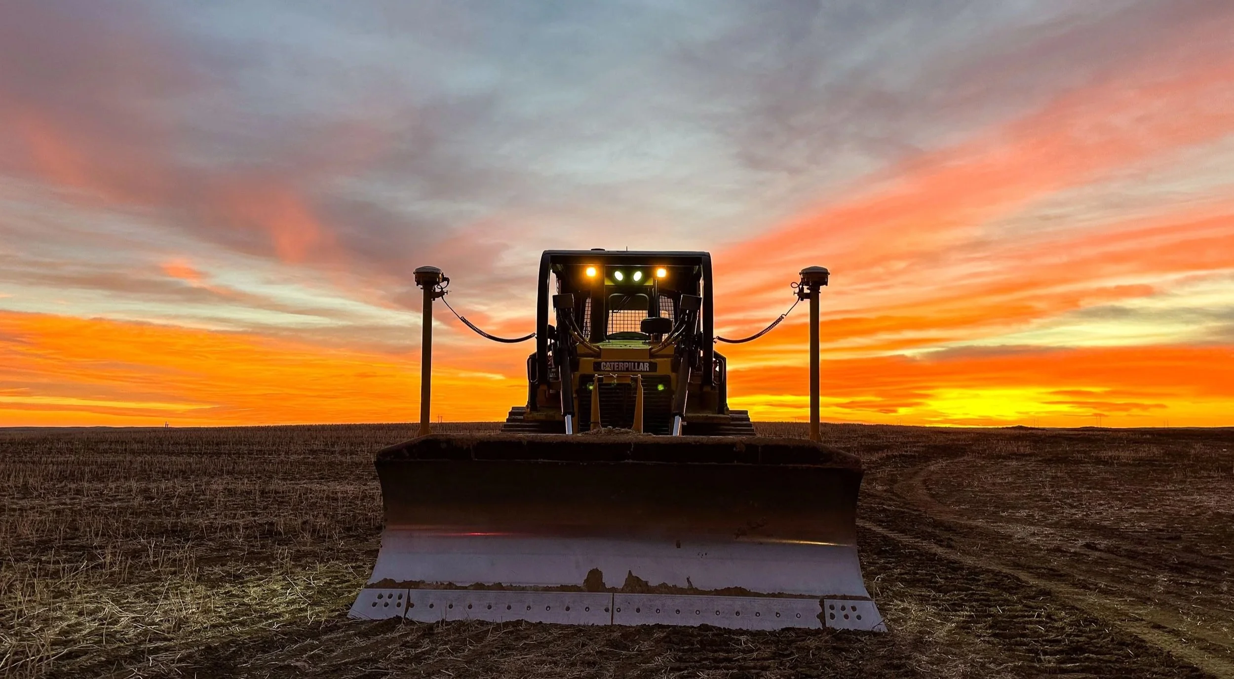 Front view of a bulldozer with Trimble GPS technology, parked on a farm field during a colorful sunset sky with clouds.