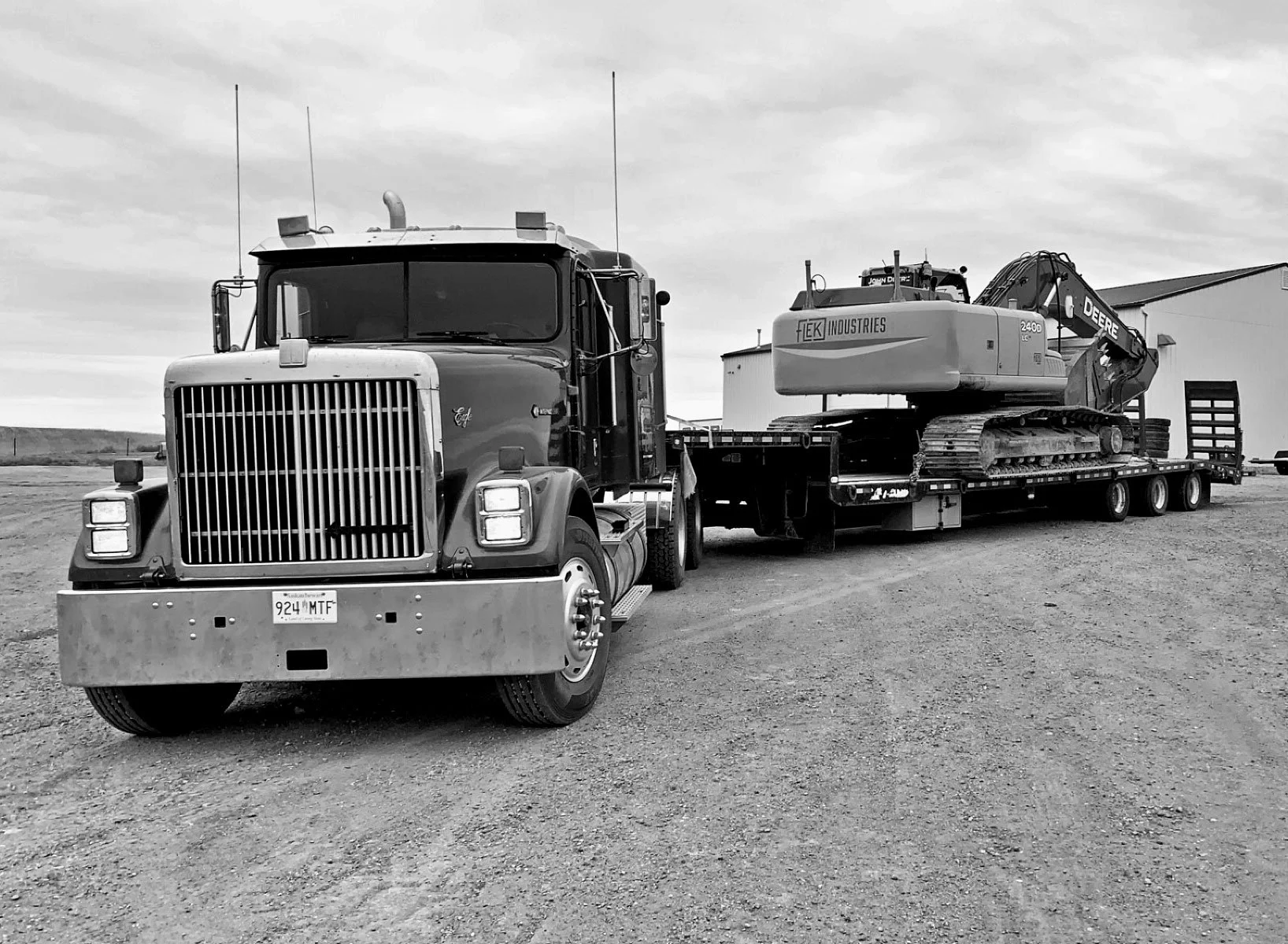 A semi-truck with a flatbed trailer carrying a large excavator, parked on a gravel surface with a building in the background.