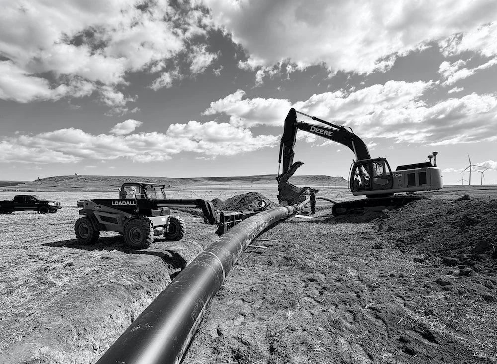 Construction scene with an excavator and a loader working on a pipeline installation in Southern Alberta with wind turbines in the background.