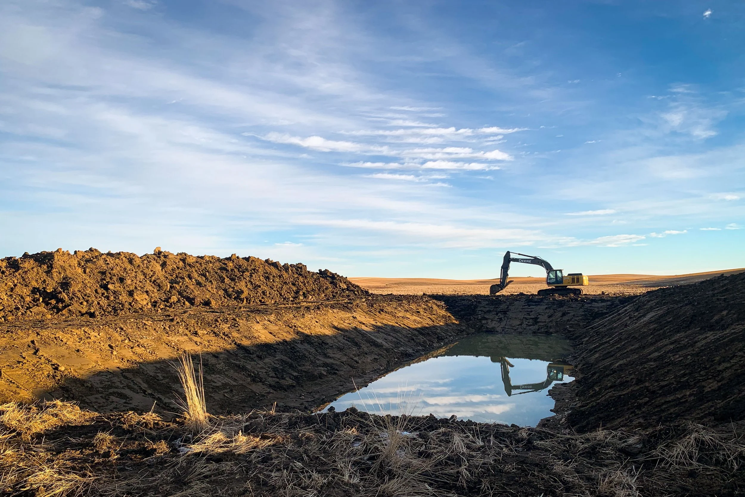 Dugout excavation being completed in rural southern Alberta, with blue sky and clouds overhead.