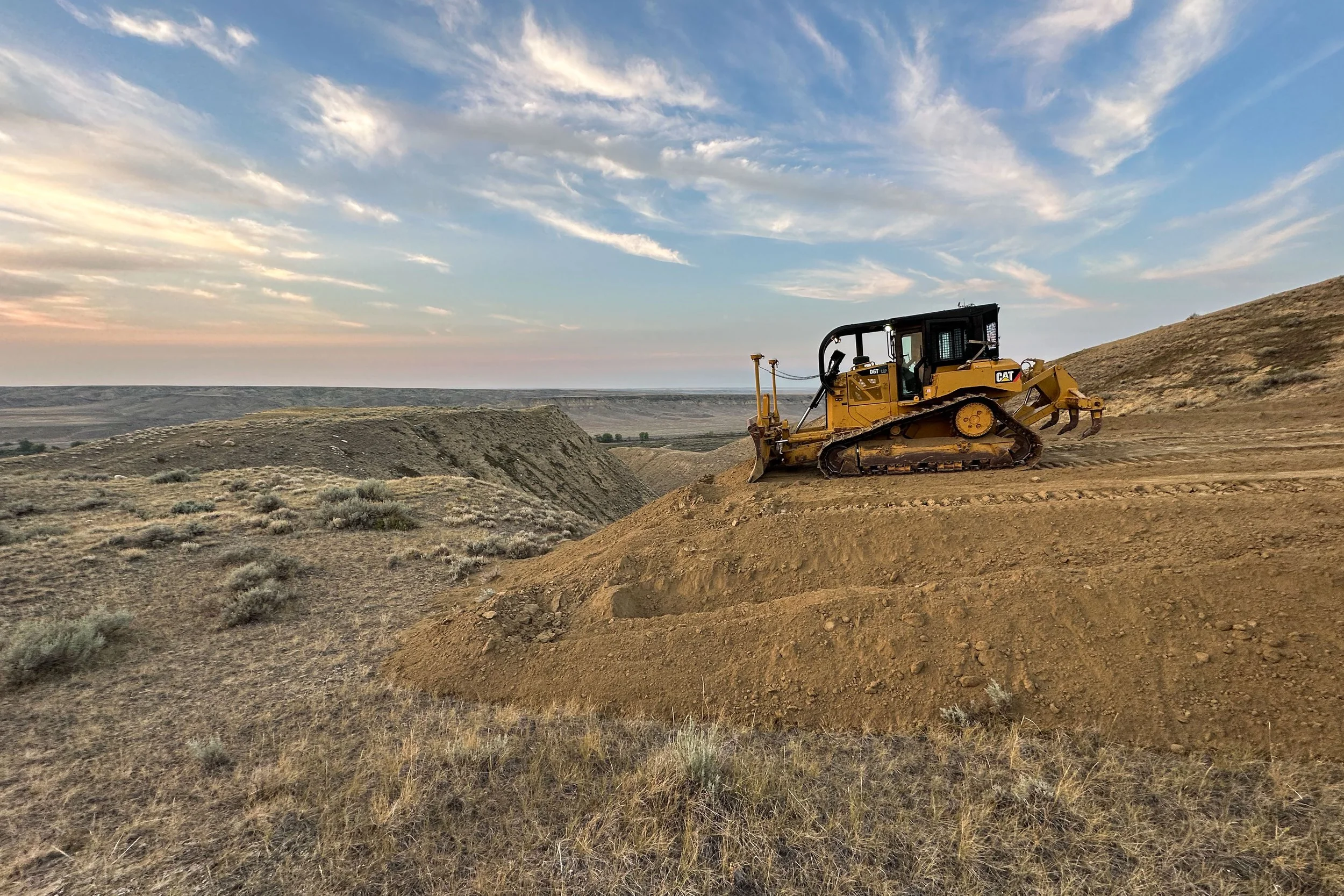 A yellow Caterpillar bulldozer with GPS Trimble working on a dirt hillside in a rural Alberta landscape under a partly cloudy sky.