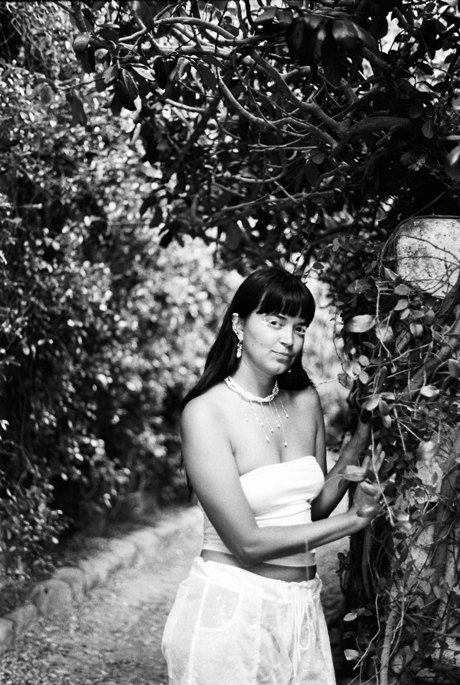 A woman with dark hair, wearing a strapless white dress and jewelry, standing outdoors among dense foliage, holding a branch with leaves.