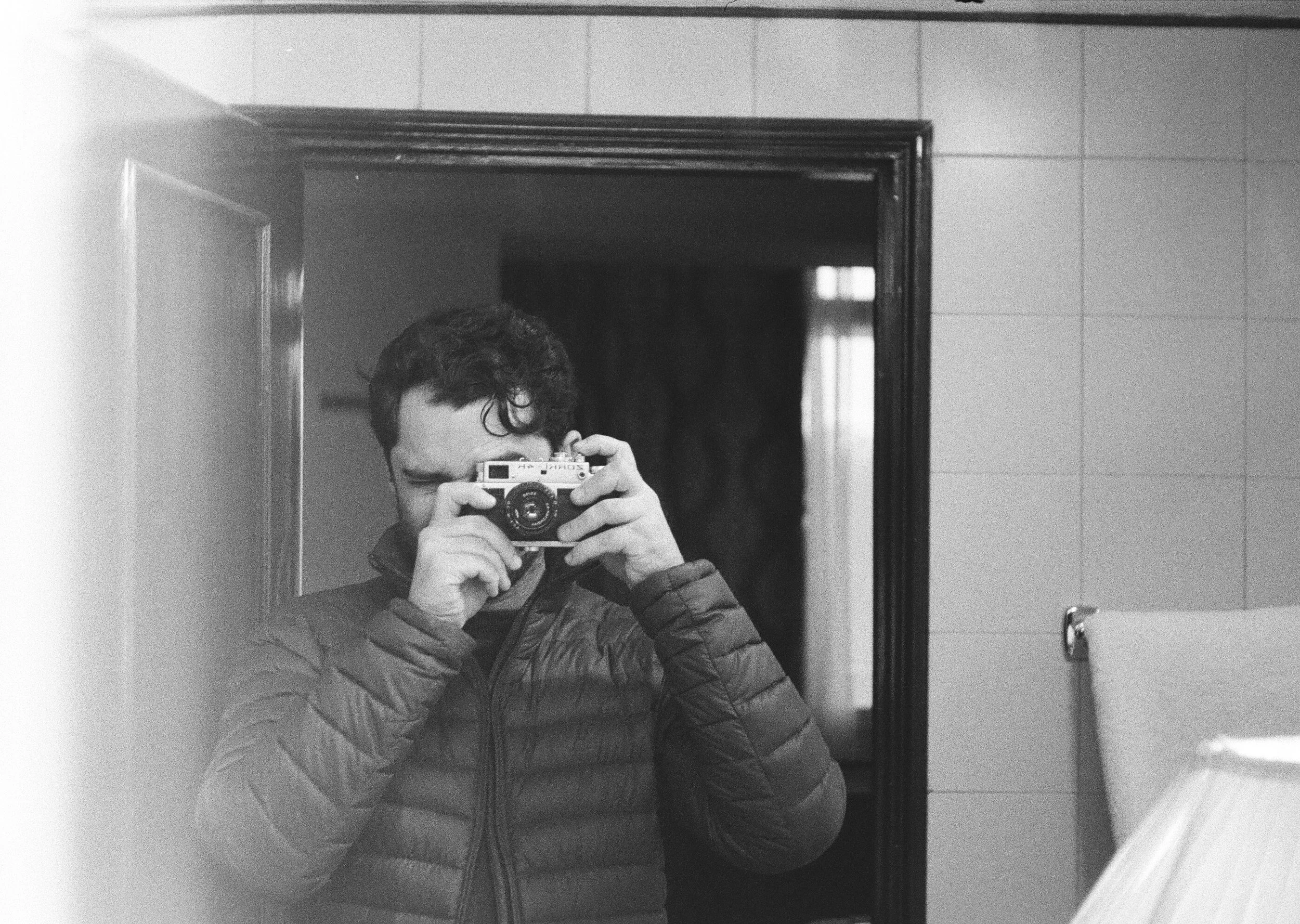 A person taking a photo of themselves in a mirror in a bathroom, holding a camera with both hands. The person has curly hair and is wearing a puffy jacket. The bathroom has tiled walls and a mirror with a dark frame.