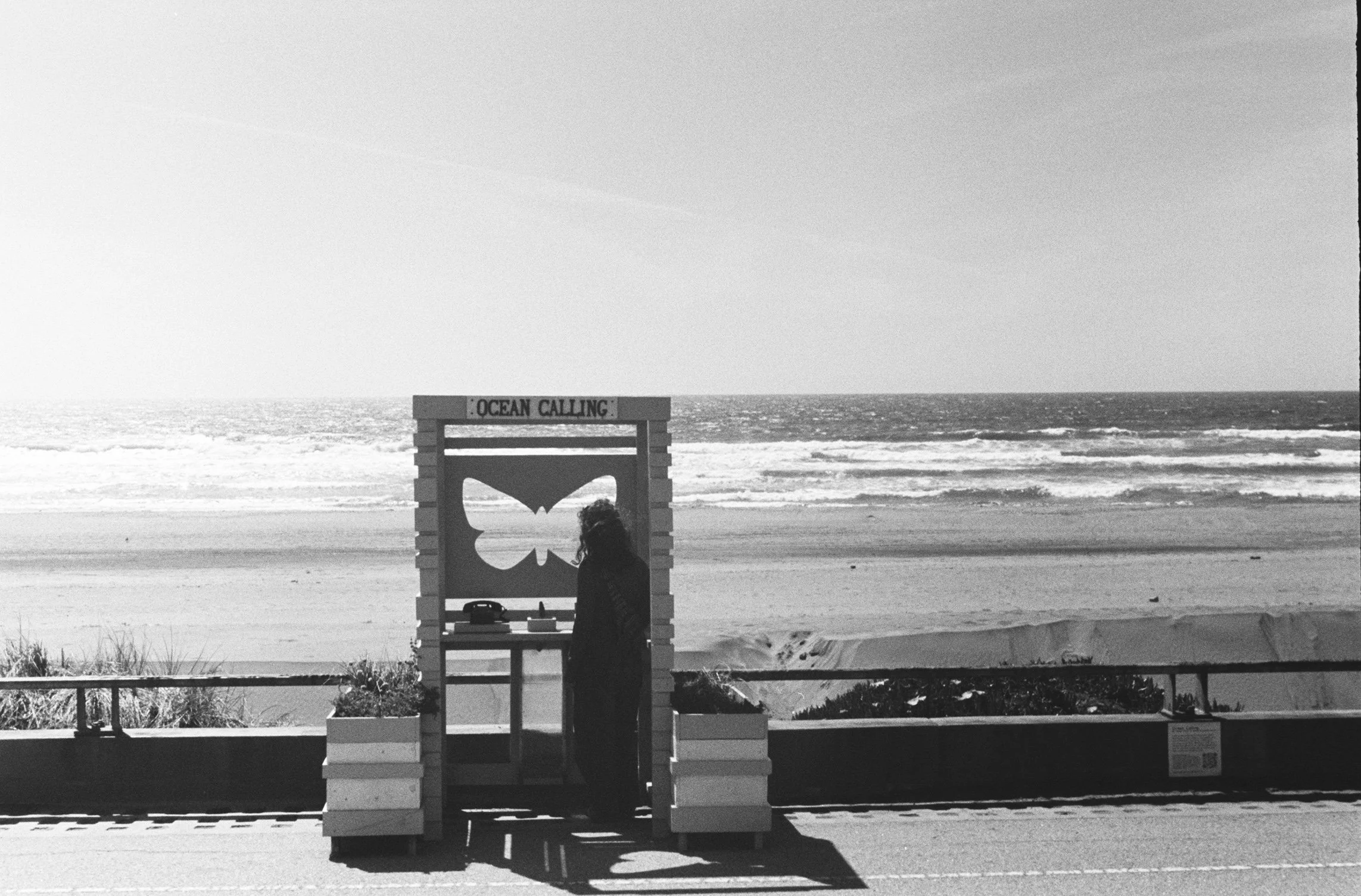 Black and white photo of a woman standing next to a small wooden cutout of a person at the beach. The cutout has the words 'OCEAN CALLING' on top. The ocean and sky are visible in the background.