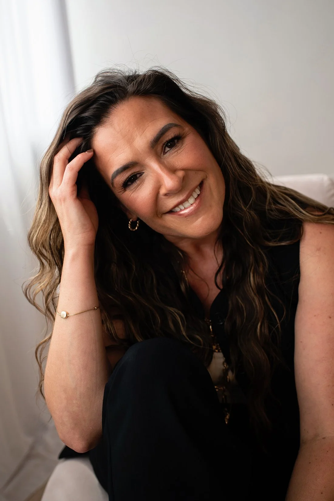 Chantal Menard smiling and touching her head, wearing a black sleeveless top, gold hoop earrings, and a gold bracelet, sitting indoors against a plain light-colored wall.