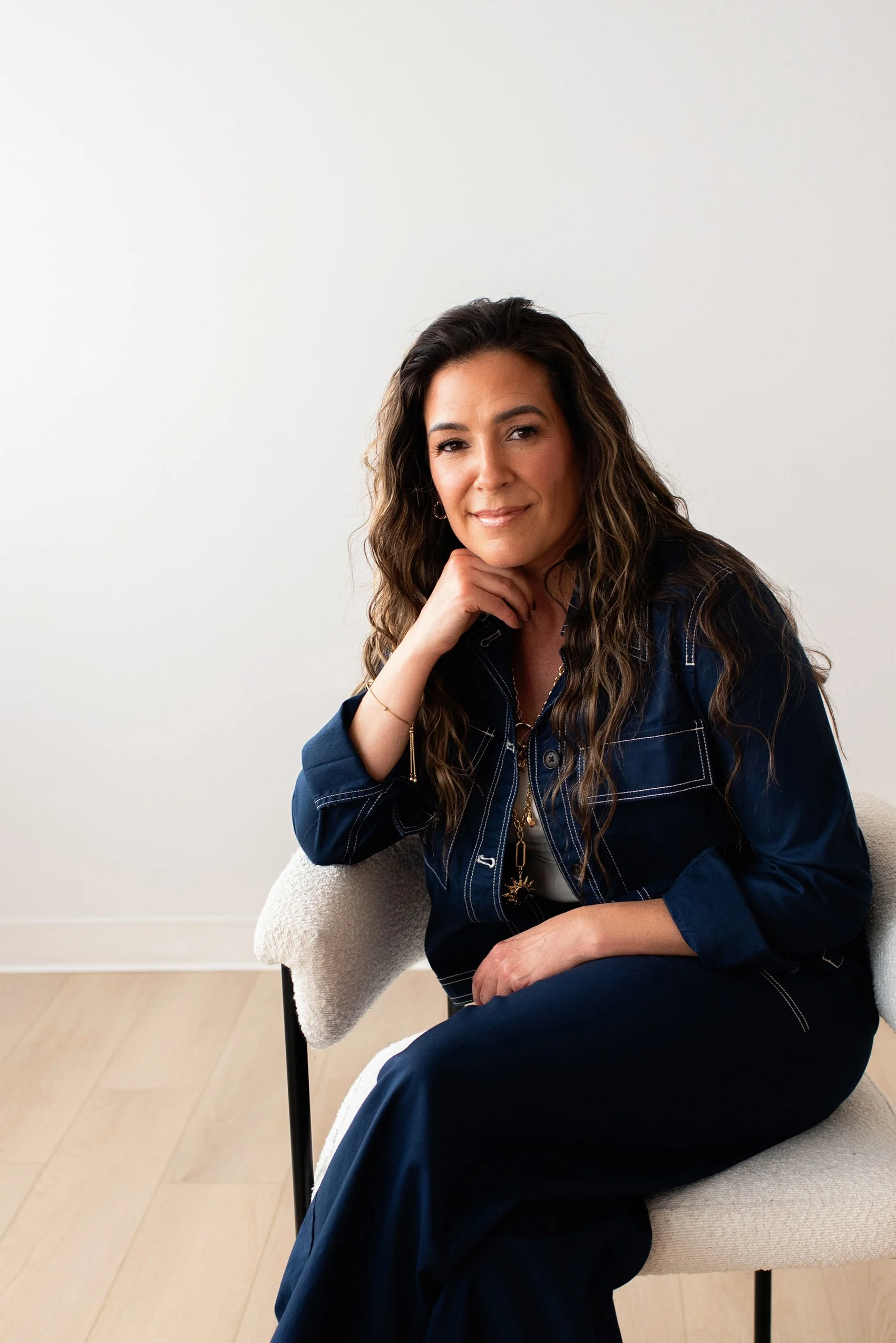 Chantal Menard sitting on a white cushioned chair, smiling with her left hand resting on her chin, wearing a dark blue denim jacket and pants, with a white top underneath, in a minimalistic room with a light-colored wooden floor and white walls.