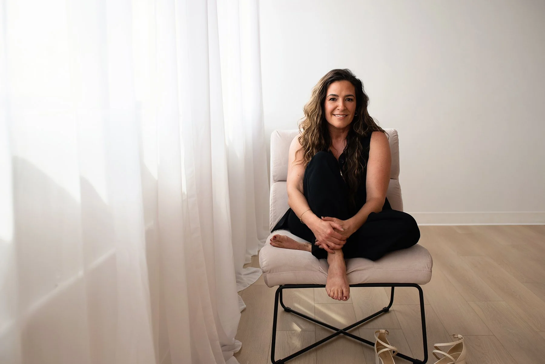 Chantal Menard sitting cross-legged on a cushioned chair in a bright room with white walls and curtains. She is smiling and wearing black clothing, with beige shoes on the floor nearby.