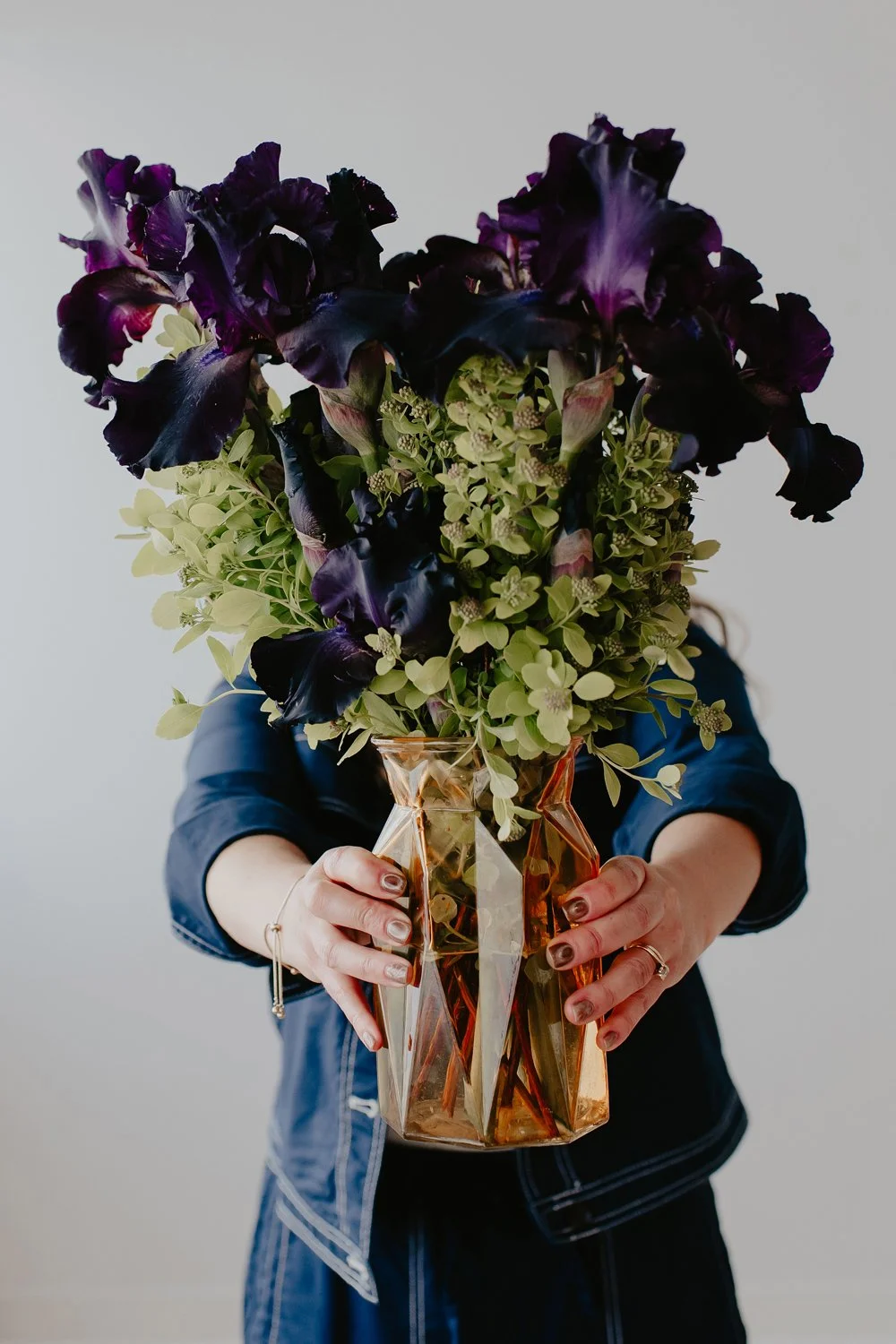 Person holding a gold-colored geometric vase filled with dark purple flowers and green foliage in front of a plain white background.