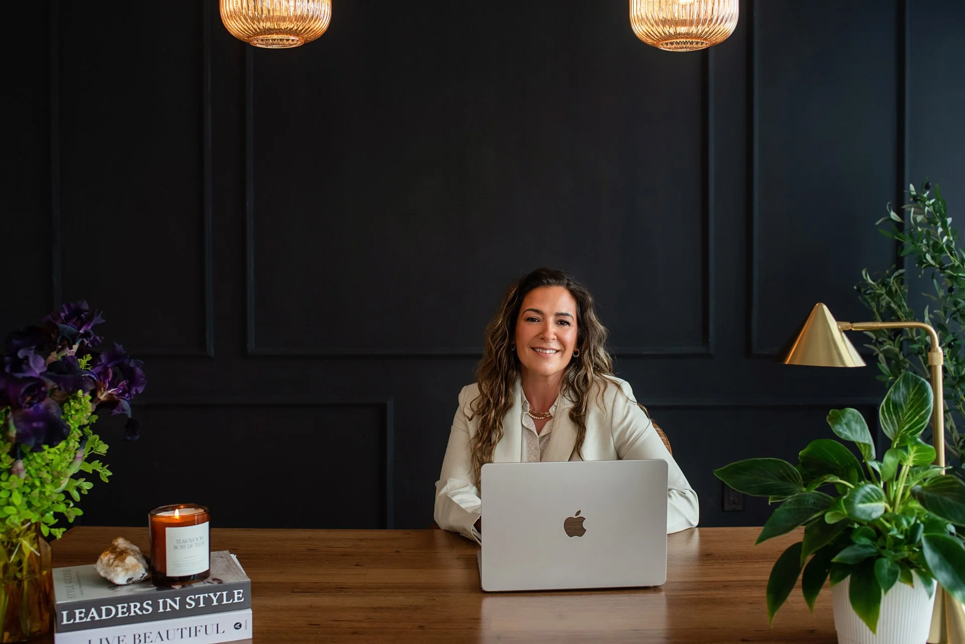 A woman with wavy brown hair and a white blazer sitting at a wooden desk with a silver MacBook. The background features dark walls, two hanging pendant lights, a gold desk lamp, and various potted plants. On the desk are a lit candle in a brown container, a book titled "LEADERS IN STYLE," and a crystal. Flowers and greenery are in the foreground.