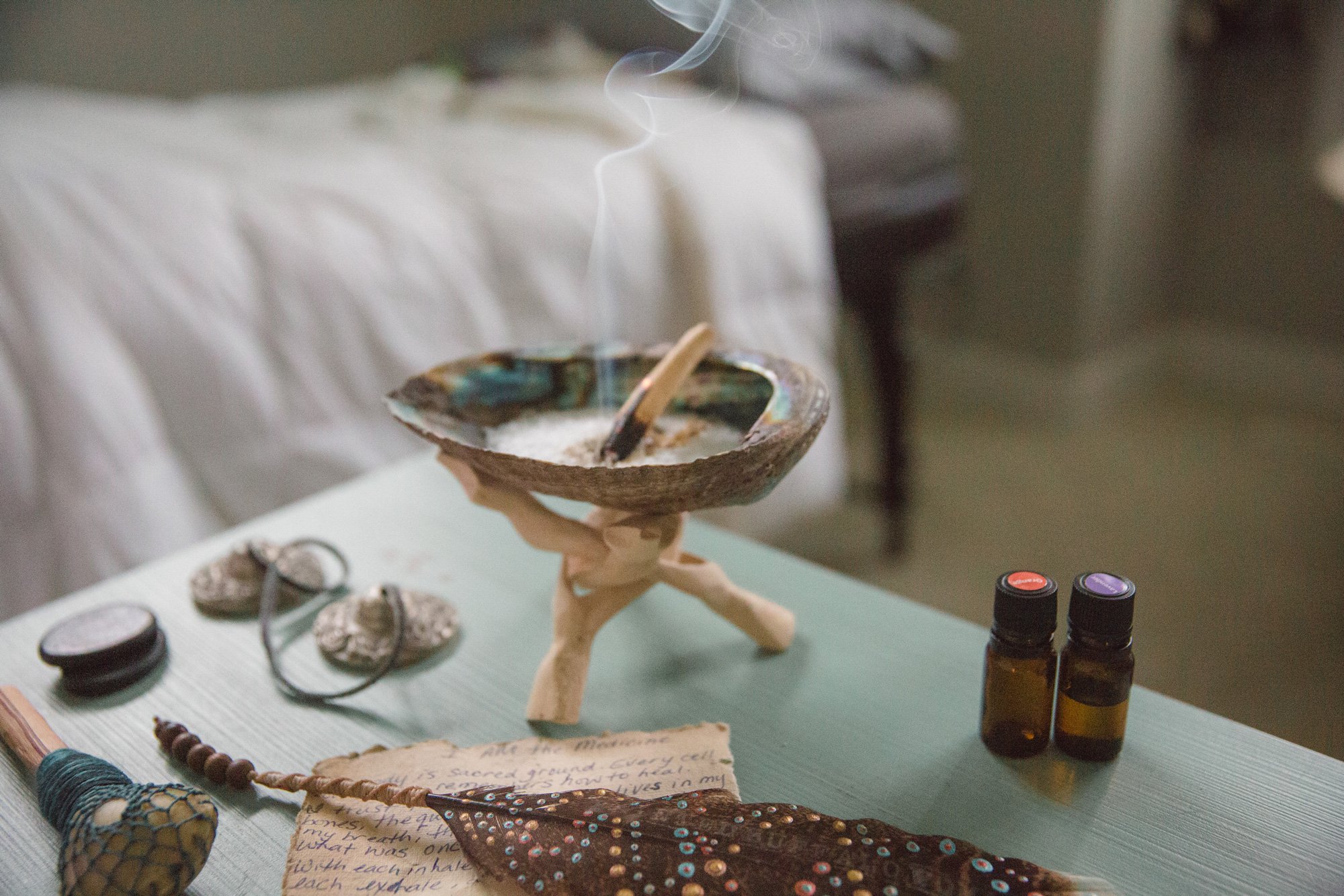 A small decorative shell holding burning incense, with white smoke curling upwards. Various small objects such as stones, bottles, and handmade craft items are arranged on a white surface underneath, likely part of a spiritual or craft display.