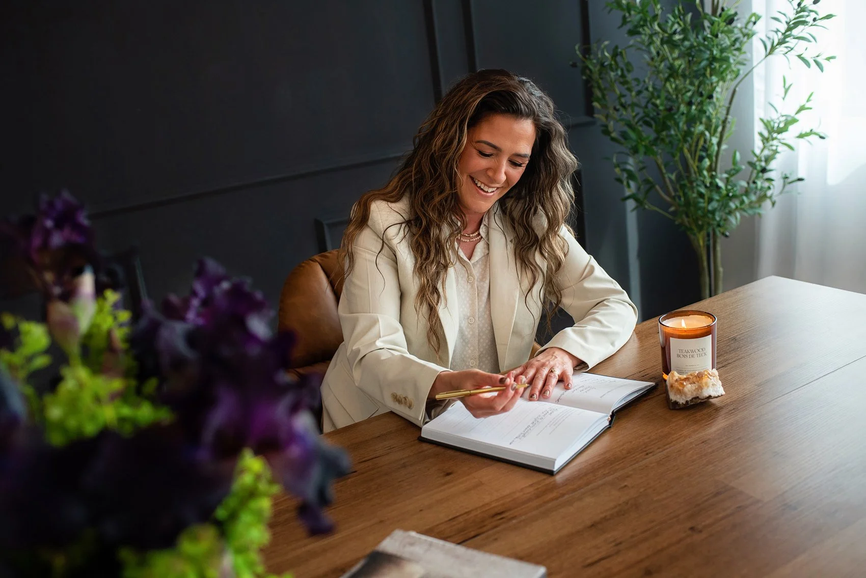 Chantal Menard in a cream blazer sitting at a wooden table, reading a book with a gold pen. There is a lit candle and a piece of mineral or crystal on the table, and potted plants in the background.