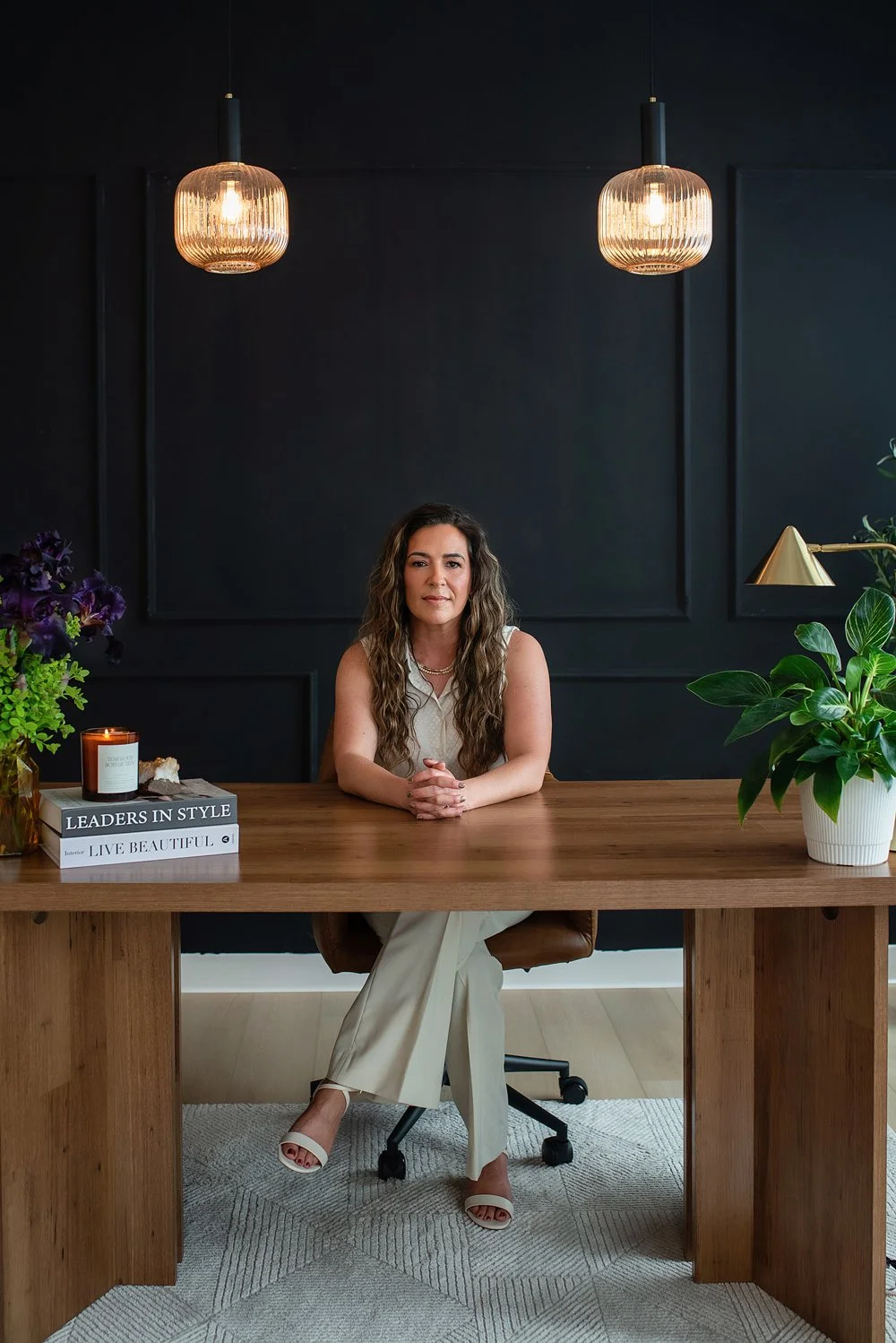 Chantal Menard sitting at a wooden desk in a modern office with dark walls, two pendant lights, a stack of books, a candle, a flower arrangement, and a large potted plant.