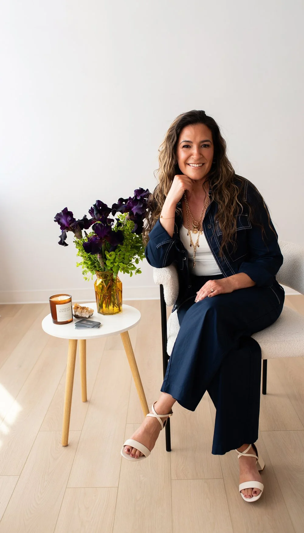 A woman with long, wavy hair, smiling and sitting in a white chair, wearing a white top, dark blue blazer, and dark blue pants, with beige high heel sandals. She is sitting next to a small white table with a yellow vase of purple and green flowers, a brown candle, a crystal, and business cards.