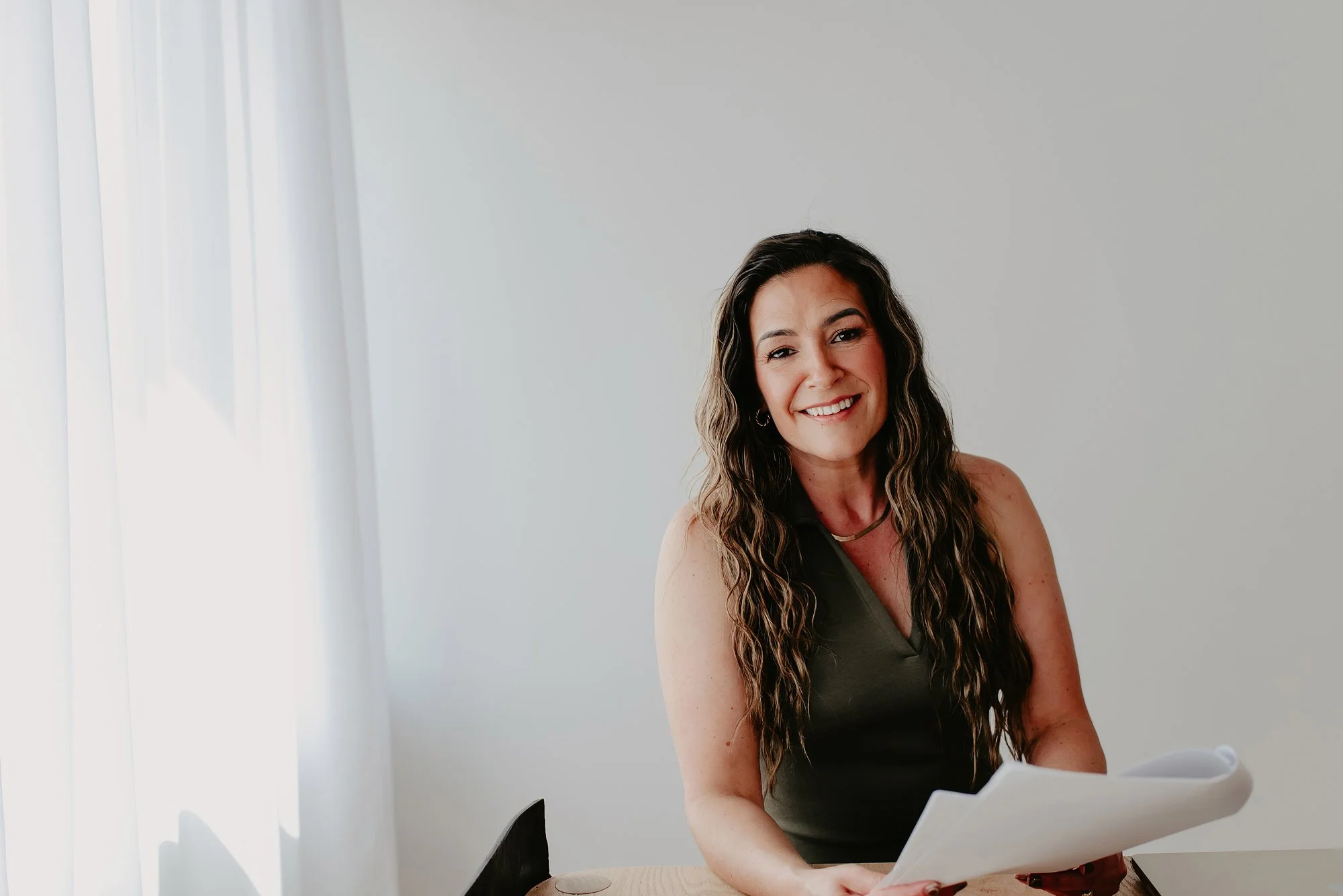Chantal Menard smiling, sitting at a desk with papers, in a room with white walls and vertical blinds.