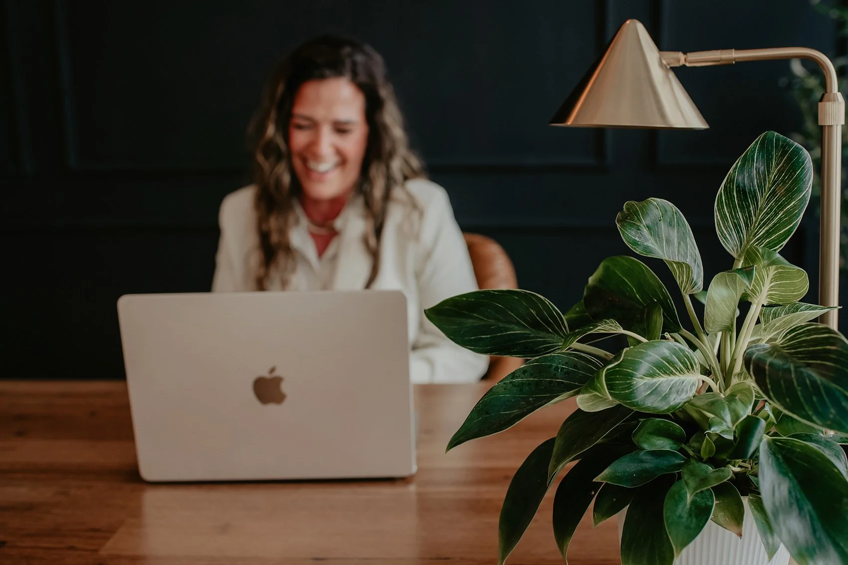 Chantal Menard smiling while working on a laptop at a desk, with a large green leafy plant and a gold desk lamp in the foreground, and a dark wall in the background.
