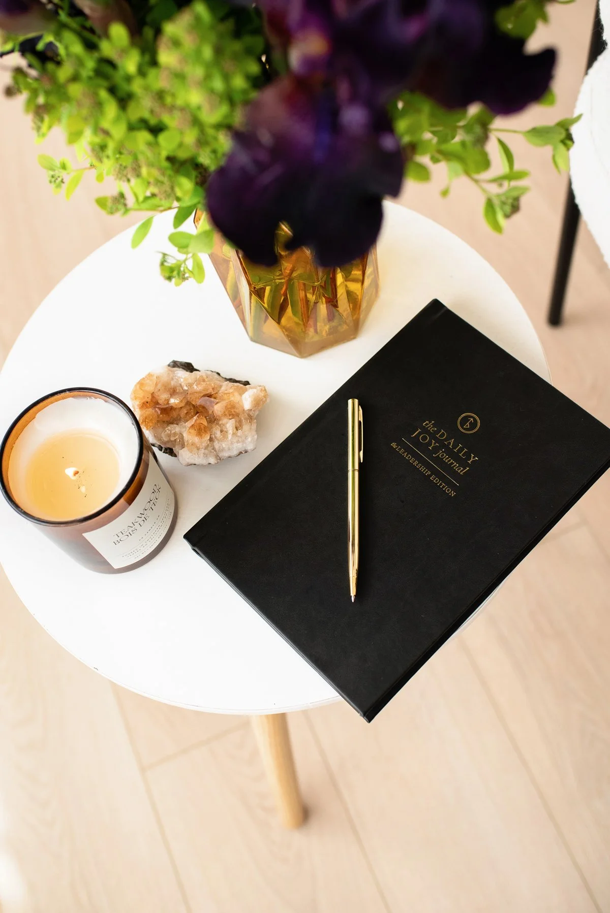 A round white table holds a black journal titled 'The Daily 10' with a gold pen on top, a lit candle in a dark amber jar, a piece of mineral or crystal, and a gold geometric vase with green and purple flowers. The table is on a light-colored wooden floor.