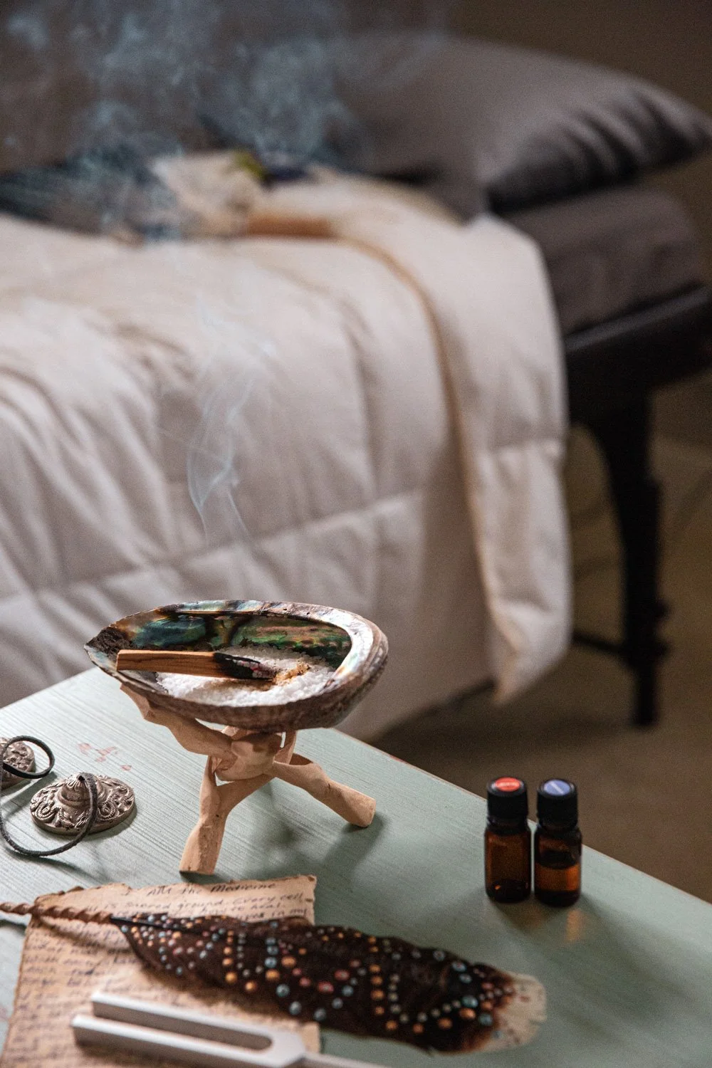 Showcase with incense, essential oils, a beaded necklace, and a shell dish on a table with a bed in the background.