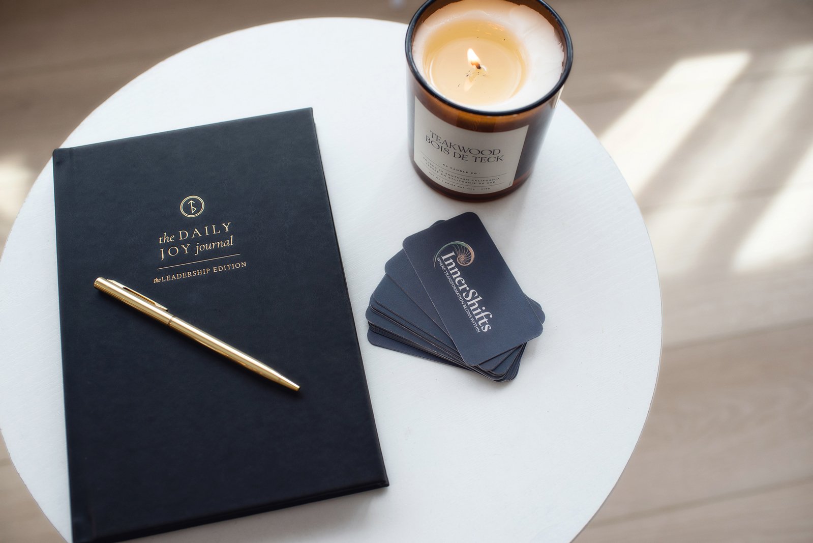 A white round table with a black journal titled 'the DAILY JOY journal, the LEADERSHIP EDITION,' a gold pen resting on the journal, a lit candle in a glass container labeled 'TEAKWOOD BOIS DE TECK,' and five black business cards that say 'Inner Shifts, LIT for Transformation, Engaging Mindful.'