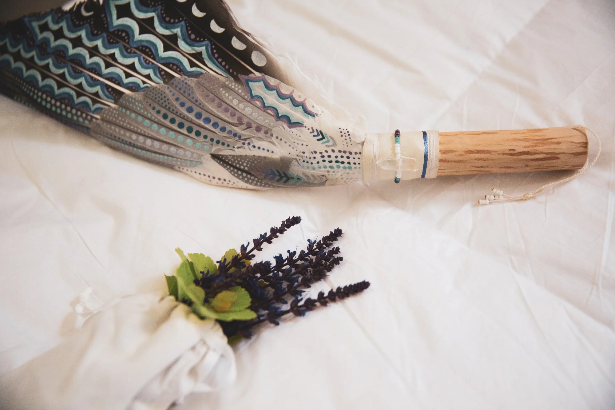 A decorative handmade umbrella with detailed patterns in blue, white, gray, and black, resting on a crumpled white surface. The umbrella handle is made of wood with a beaded decoration near the top. There are sprigs of dried lavender and some green fabric leaves placed near the umbrella.