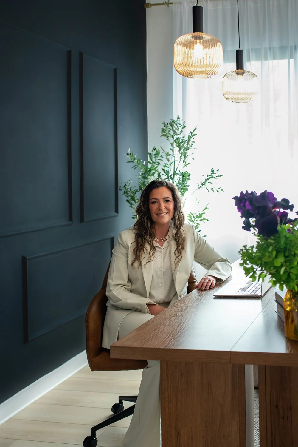 Chantal Menard in a cream suit sitting at a wooden desk in a well-lit office, with a laptop and purple flowers on the desk, and green plants behind her.