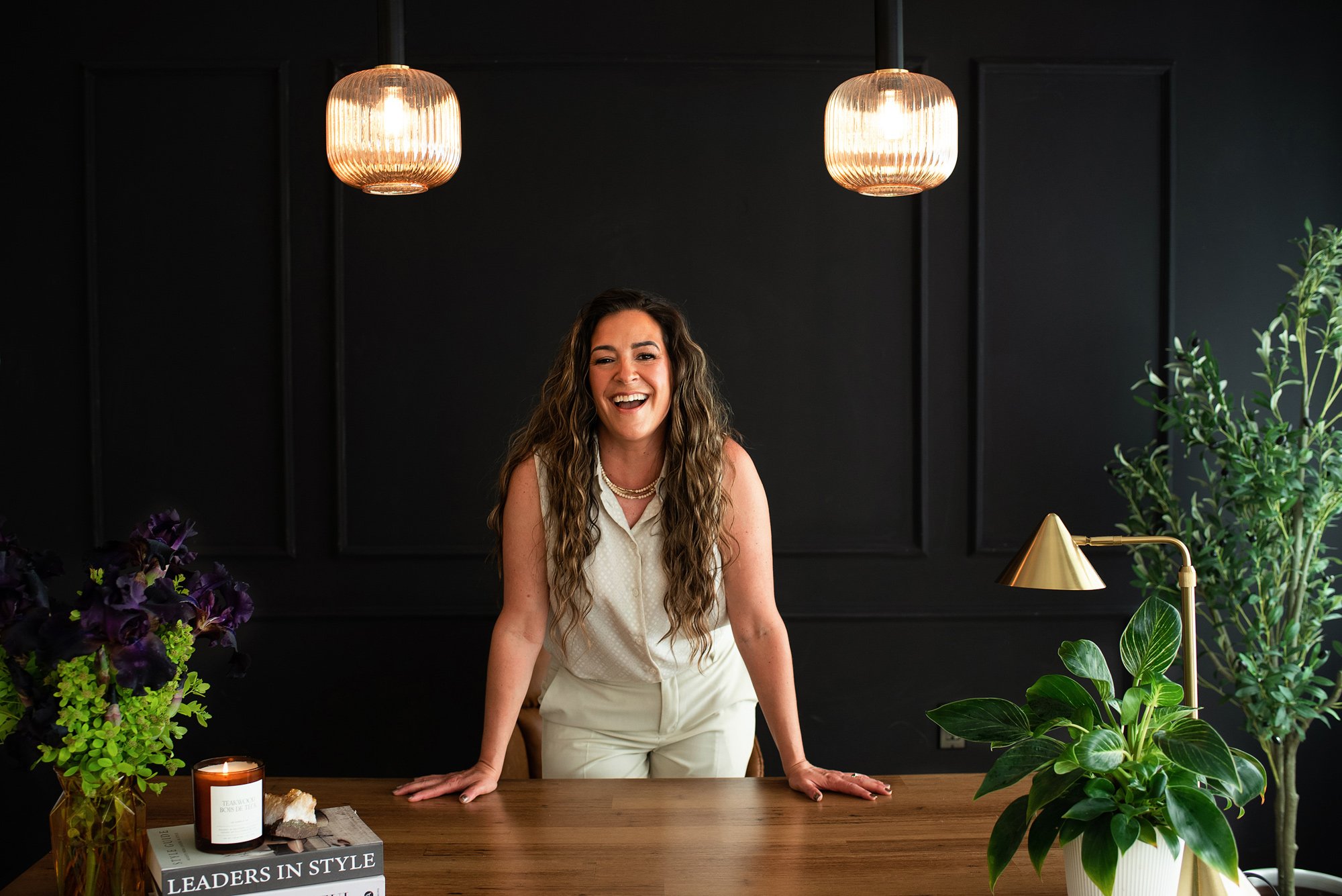 A woman with long curly hair, wearing a white sleeveless blouse and cream pants, is leaning forward on a wooden table, smiling. The background is black with two hanging pendant lights above her, and there are green plants and purple flowers on the table. A candle and a magazine are also on the table, with a book titled 'LEADERS IN STYLE' visible in the foreground.