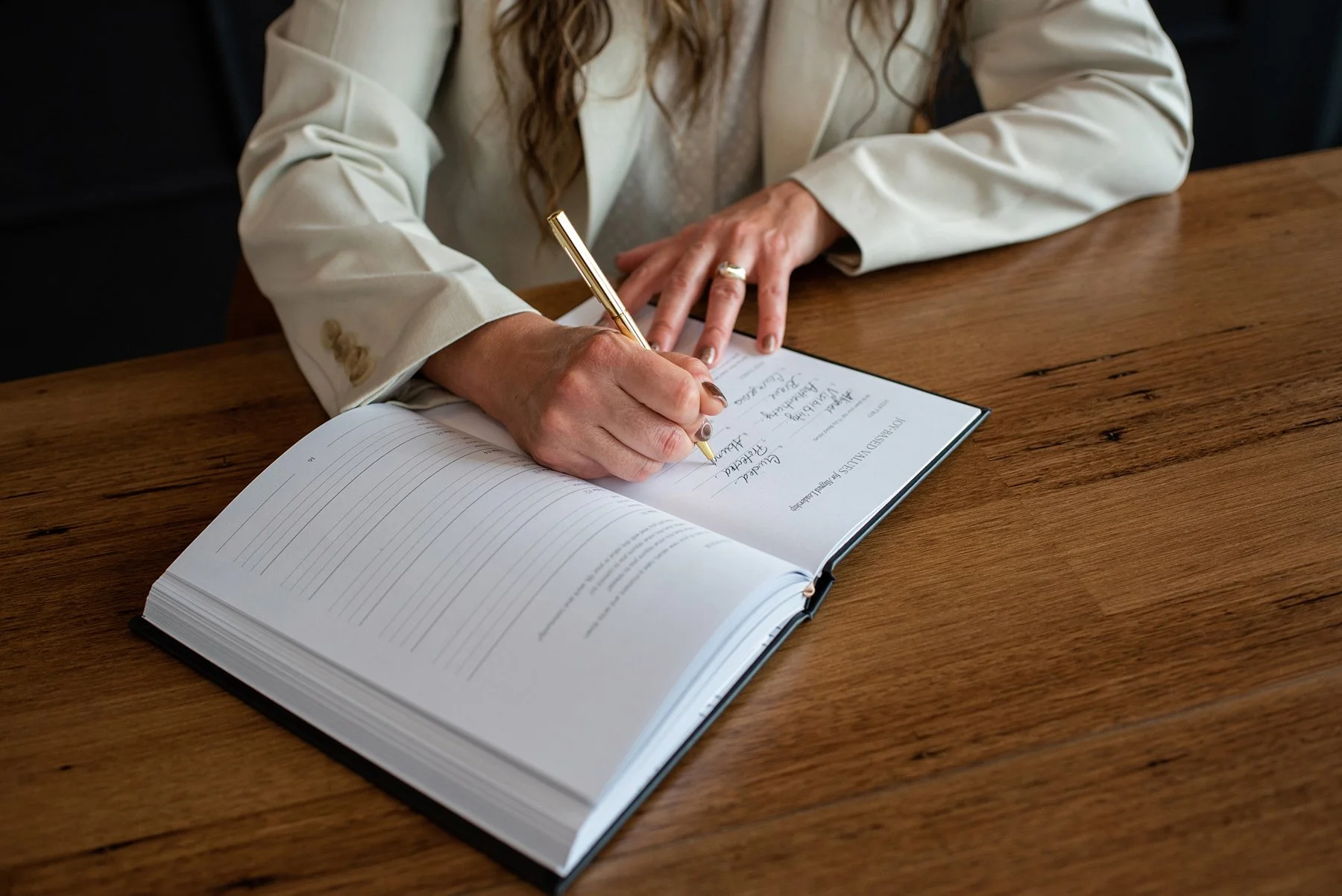 Chantal Menard wearing a beige blazer and gold ring writing in a guestbook or journal with a gold pen, on a wooden table.
