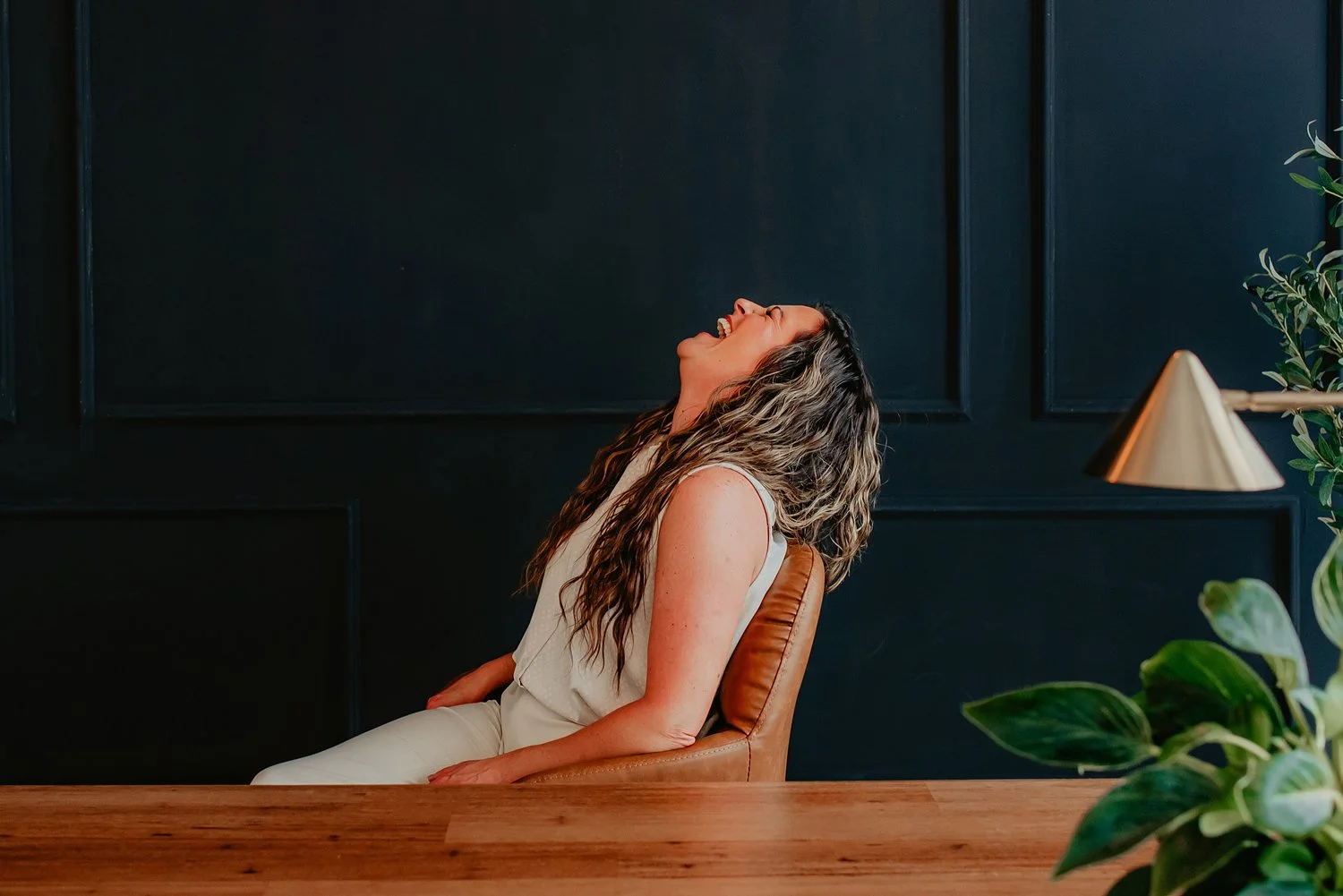 Chantal Menard sitting on a tan chair, laughing with her head tilted back. She is wearing a white sleeveless outfit. The background features a dark wall with decorative panels, a wooden table, and a green plant on the right side with a gold lamp.