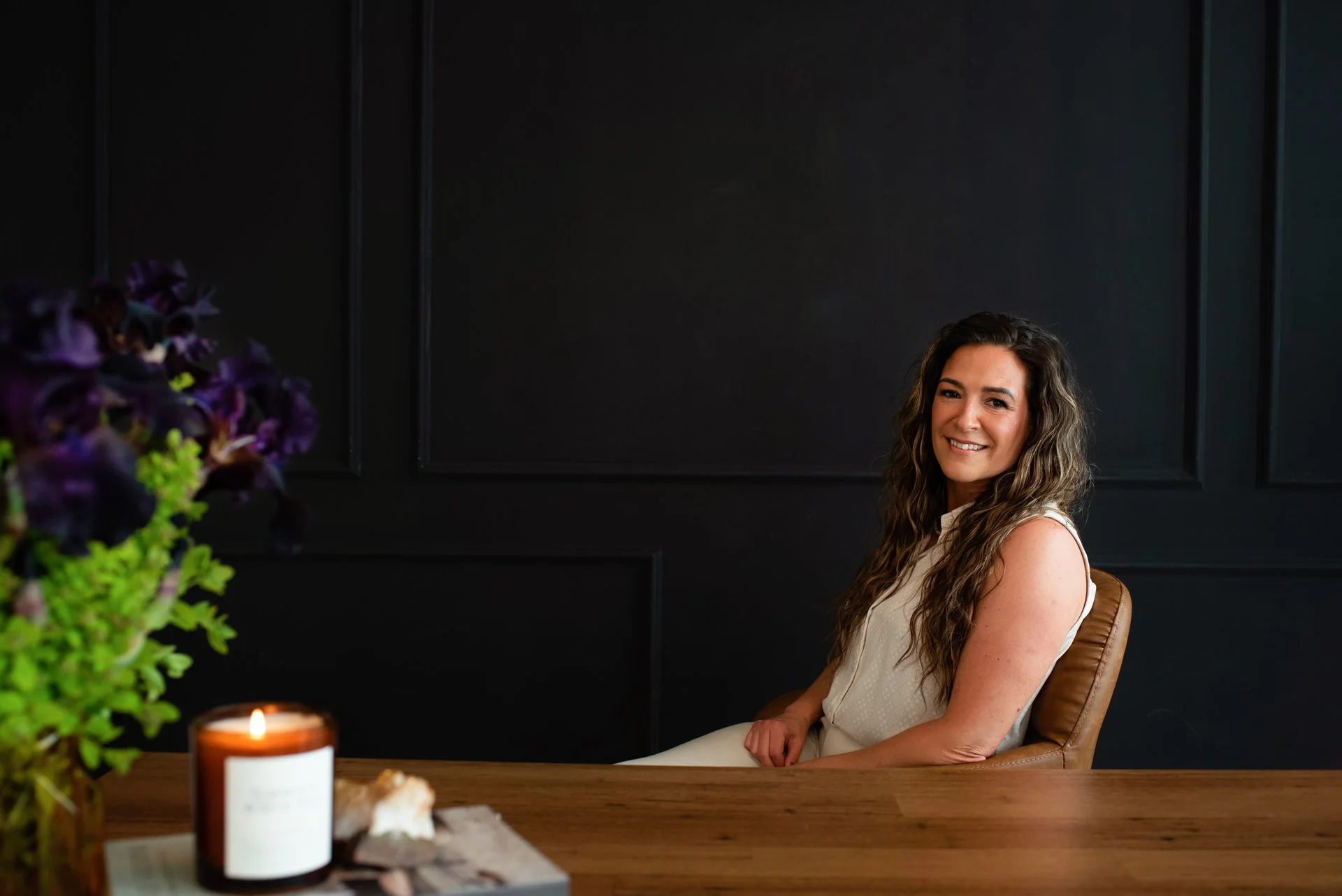 Chantal Menard smiling while sitting in a brown leather chair at a wooden table, with purple and green flowers and a lit candle in a brown container in the foreground, and a black wall with decorative panels in the background.