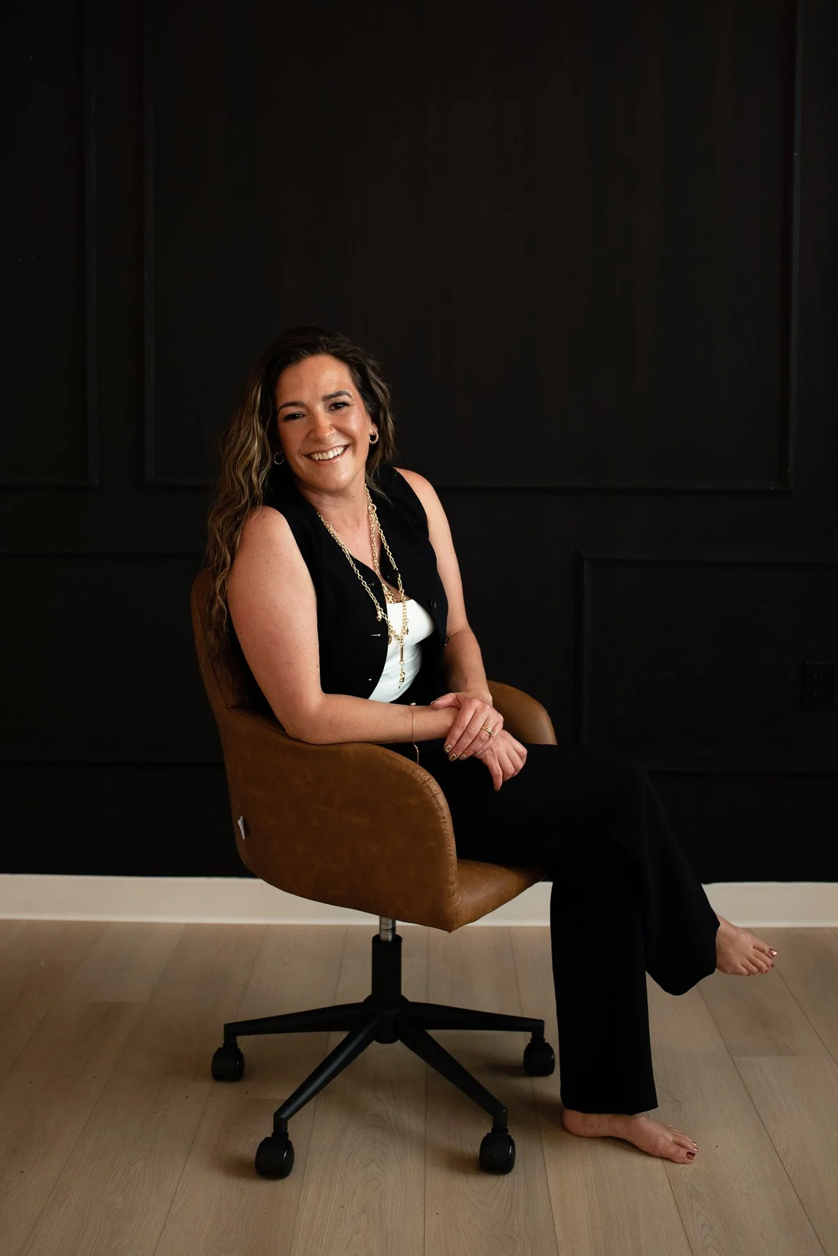 Chantal Menard sitting on a brown office chair in front of a dark wall, smiling at the camera, wearing a black sleeveless vest over a white top, black pants, and jewelry.