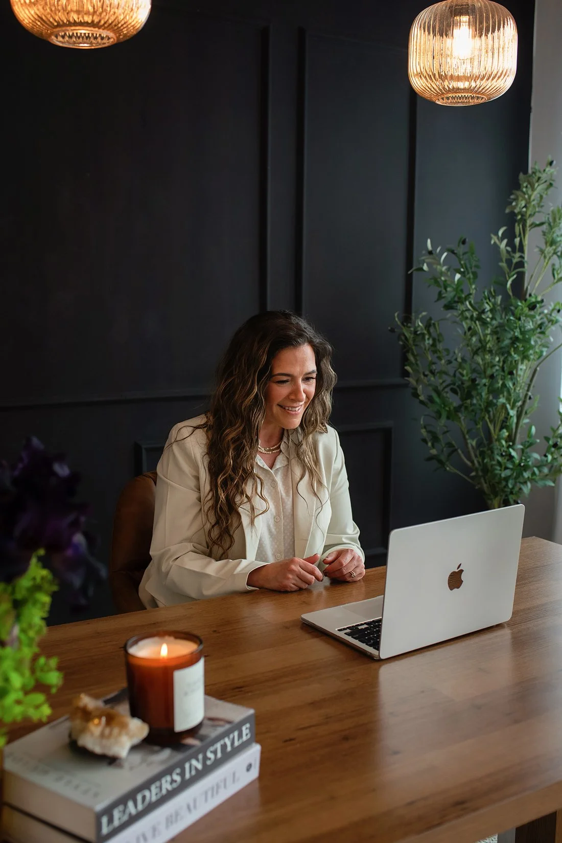 Chantal Menard smiling while sitting at a wooden table, working on a laptop in a modern, dark-walled room with a large green plant, a lit candle, and a stack of books.