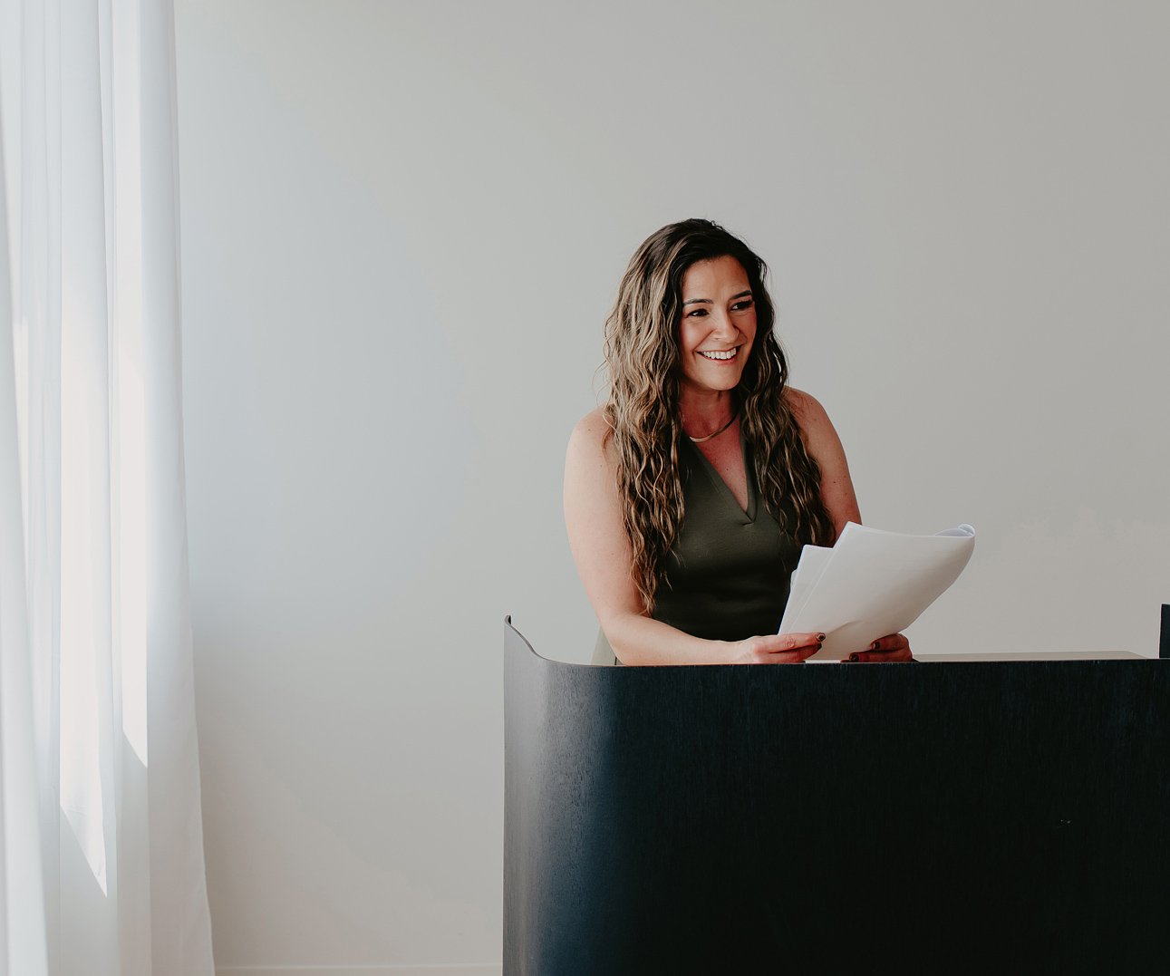 Chantal Menard smiling and holding papers, standing behind a black reception desk in a bright room with white walls and a window with sheer curtains.