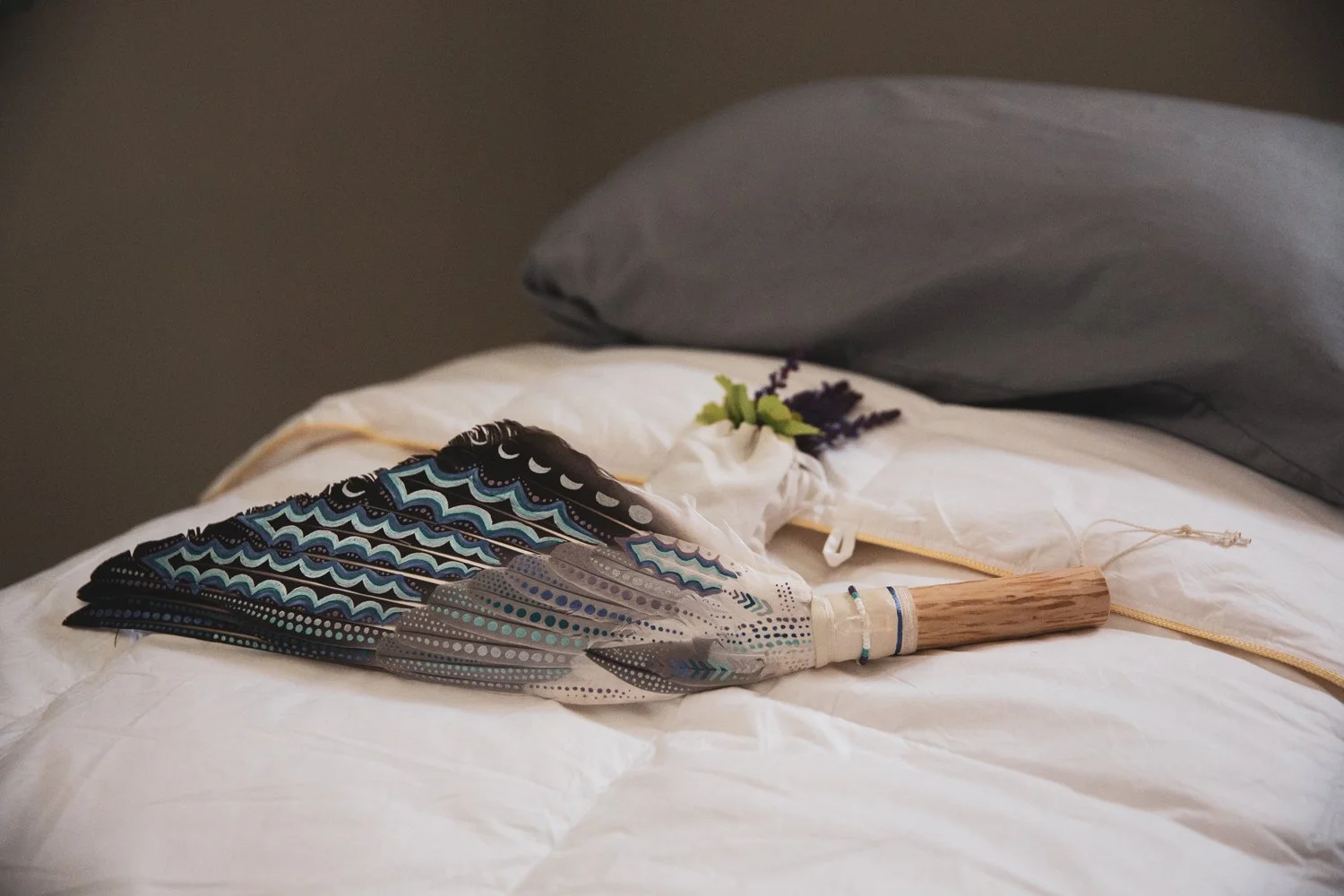 A decorative feather fan with black, white, and blue feathers and a wooden handle, resting on a white bed with a gray pillow in the background.