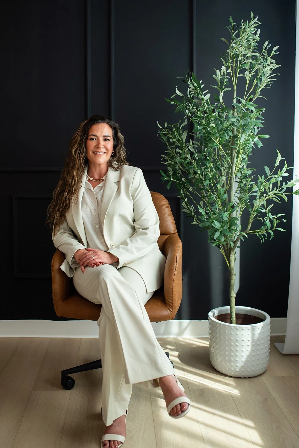 Chantal Menard dressed in a white suit, sitting on a brown leather office chair in front of a dark wall with a large green plant in a white pot beside her.