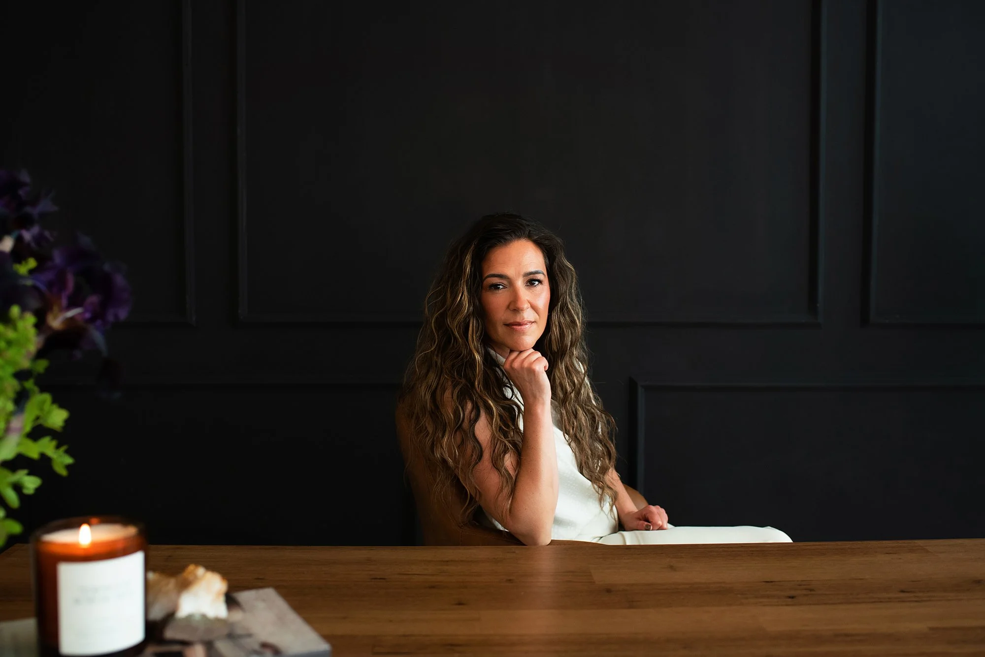Chantal Menard sitting at a wooden table in front of a black wall, with a candle and flowers partially visible in the foreground.