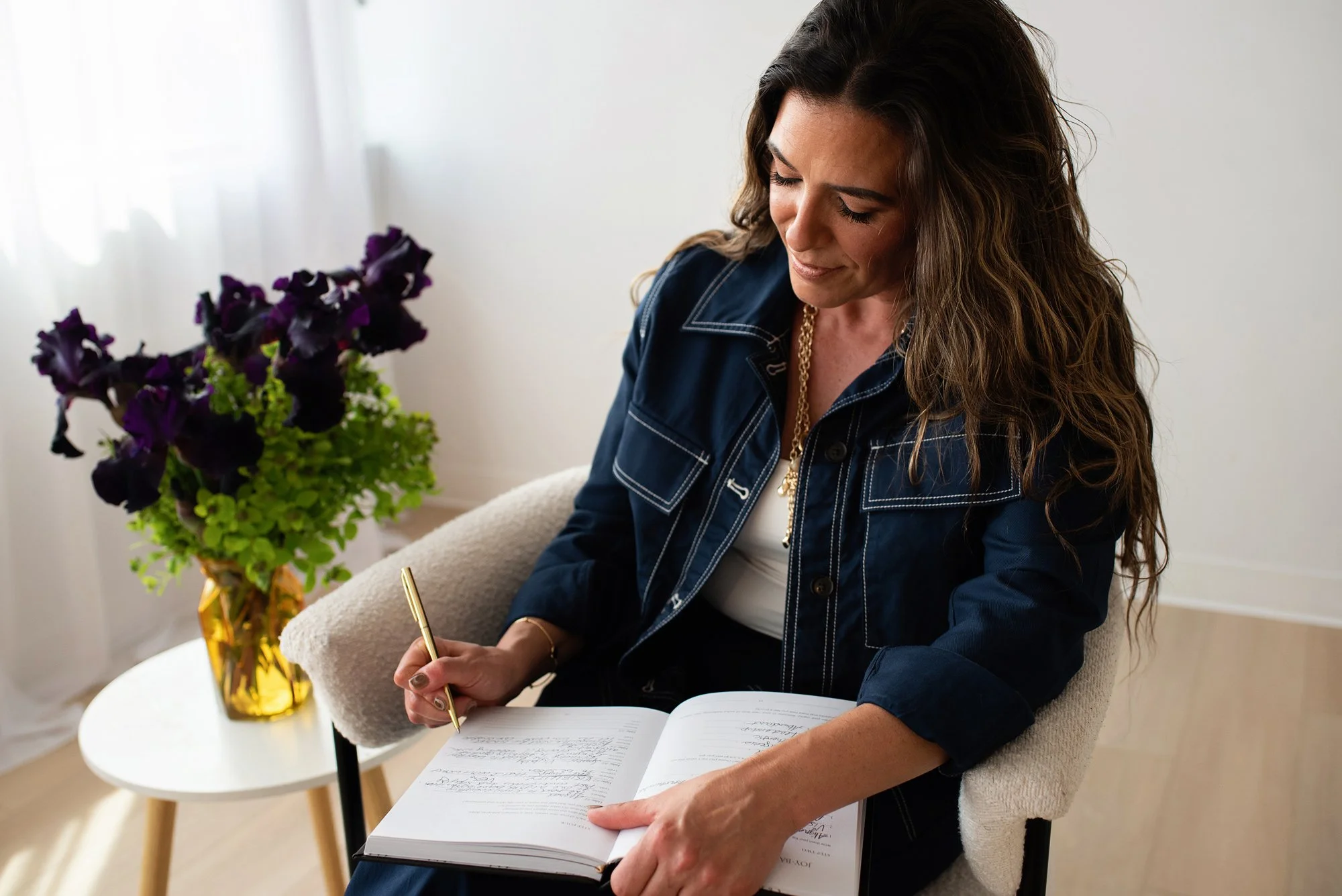 A woman with long, wavy brown hair, wearing a denim jacket with white stitching and a gold chain necklace, sits in a cozy room with natural light. She is smiling gently, writing in a notebook with a gold pen, seated in a beige armchair with a soft cushion. On a small white table next to her, there is a vibrant yellow vase filled with purple and green flowers, adding a touch of color to the space.