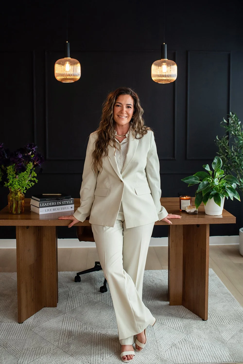 Chantal Menard standing in an office with a dark wall background, wooden desk, green plants, books, and hanging pendant lights.