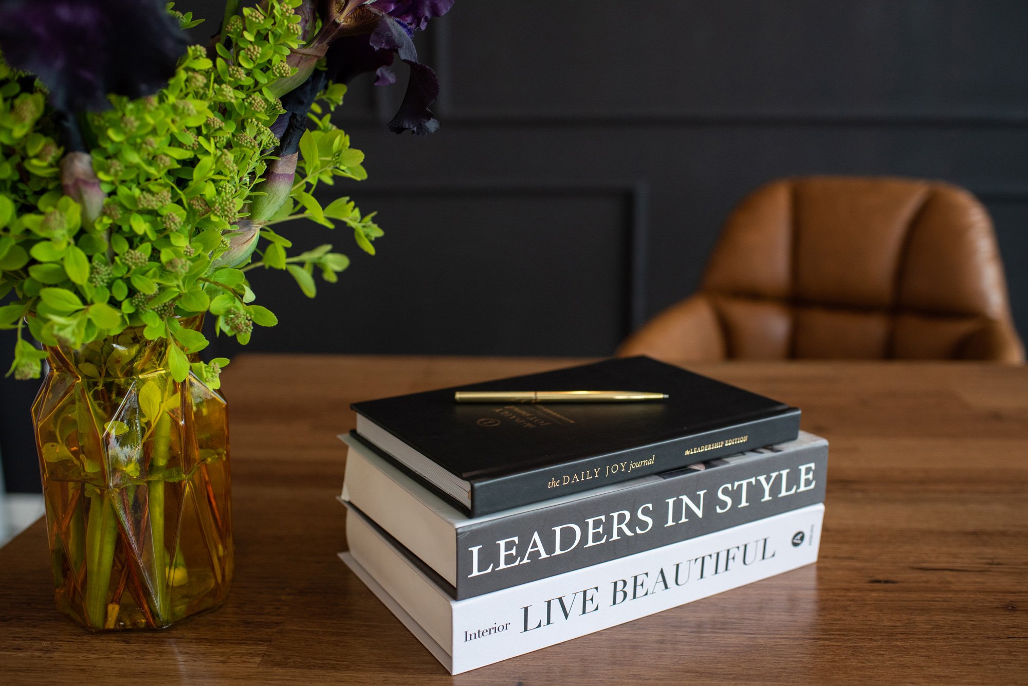 A wooden table with a yellow transparent vase holding green and purple flowers on the left, a stack of five hardcover books with titles related to leadership, interior design, and beauty in the center, and a brown leather chair in the background against a dark wall.