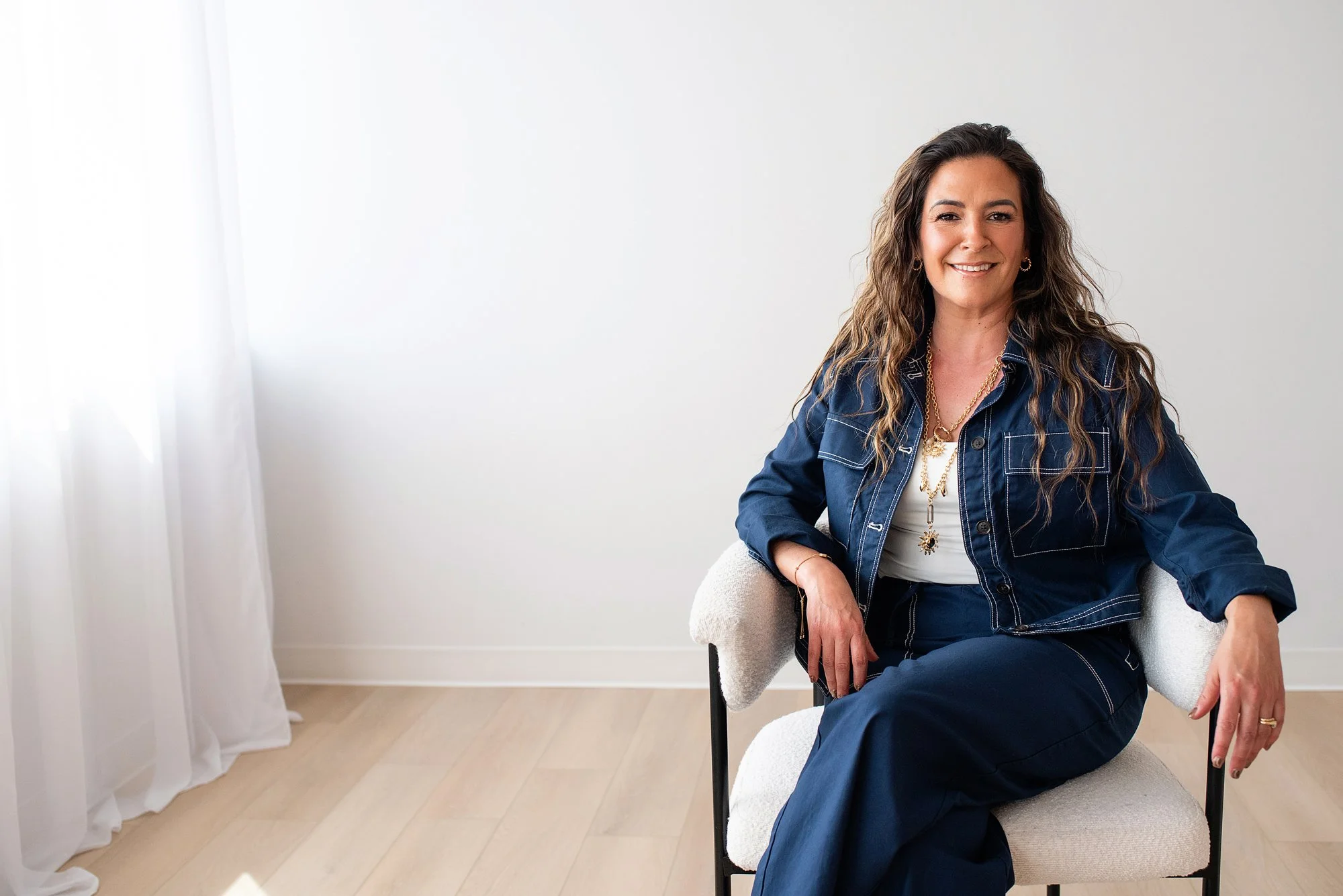 Chantal Menard smiling, sitting on a light-colored armchair in a room with white walls and a window with sheer curtains.